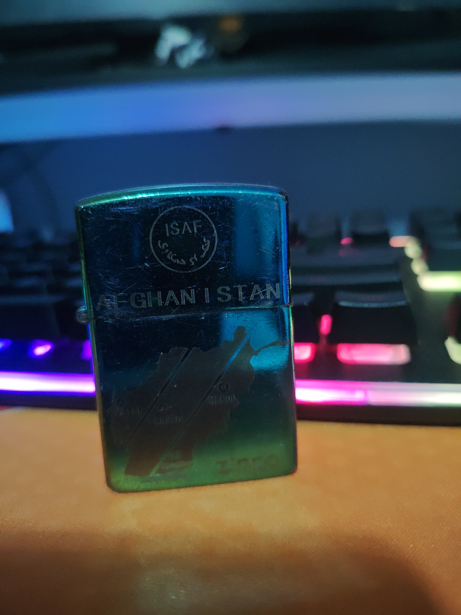 A metallic, Zippo-style lighter with an iridescent blue-green finish is centered in the foreground, resting on an orange, textured surface, likely a desk or mousepad. The lighter shows signs of wear, with numerous scratches across its surface.
The front of the lighter features several engraved details. At the top, a circular emblem contains the text "ISAF" in English, along with some script in Arabic characters beneath it. Directly below this emblem, the word "AFGHANISTAN" is engraved in prominent capital letters. Further down the lighter, an outline map of Afghanistan is etched, with several cities marked within it, including "Kabul," "Herat," and "Kandahar." The "Zippo" logo is partially visible towards the bottom right of the map.
In the blurred background, a computer keyboard with multi-colored RGB backlighting is visible, displaying hues of blue, purple, pink, and green. The lighting in the scene is artificial, emanating from the keyboard and possibly other sources, creating reflections on the lighter's shiny surface. The overall ambiance suggests an indoor setting, possibly at night or in a dimly lit room. No people or specific activity are depicted.