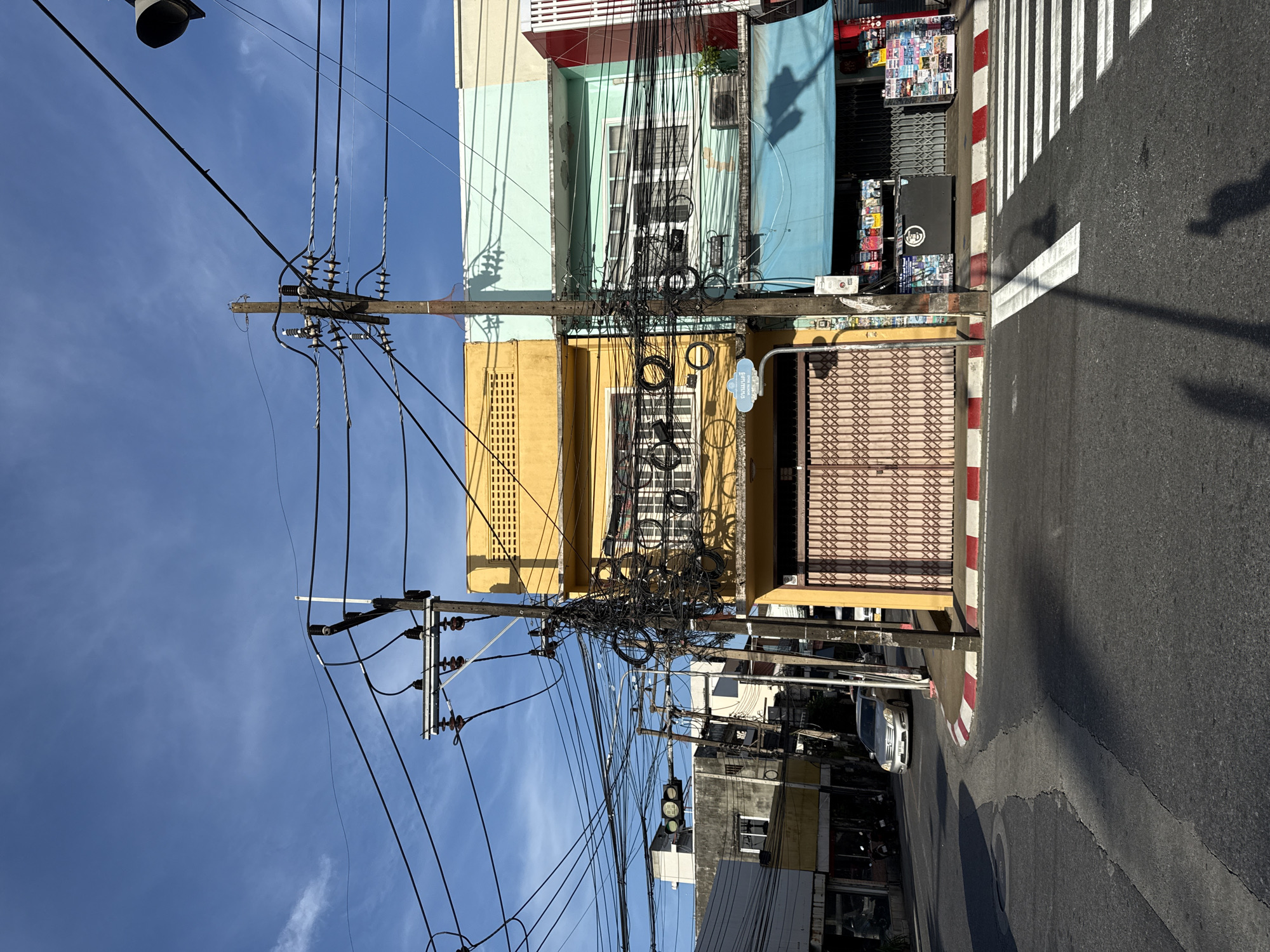 A street-level, wide-angle, outdoor photograph shows a clear blue sky with thin wisps of clouds on a sunny day. The scene captures a junction in Phuket City Municipality, Thailand, characterized by a complex and dense network of overhead electrical wires and utility poles.
In the foreground, a paved road features a pedestrian crosswalk with prominent white stripes. A shadow of a person holding a phone and another indistinct shadow are cast on the road. To the right, the white and red striped curb of the sidewalk is visible.
Dominating the mid-ground are two connected buildings. The building on the left is painted bright yellow with white-framed windows on the upper floor and a closed metallic roller shutter in a light pink color on the ground floor. A street sign, with Thai script, is mounted on a pole in front of this building. The building to its right is painted in alternating pastel colors of light green and pale blue, also with white-framed windows and air conditioning units visible. The ground floor of this building has a blue awning extending over a shopfront displaying various items on shelves.
Several utility poles, heavily laden with tangled electrical wires, are positioned throughout the scene. Traffic lights with green and red signals are visible to the left, indicating an intersection. A silver car is driving on the road in the background. The overall impression is one of a vibrant, busy urban environment with a high density of infrastructure.