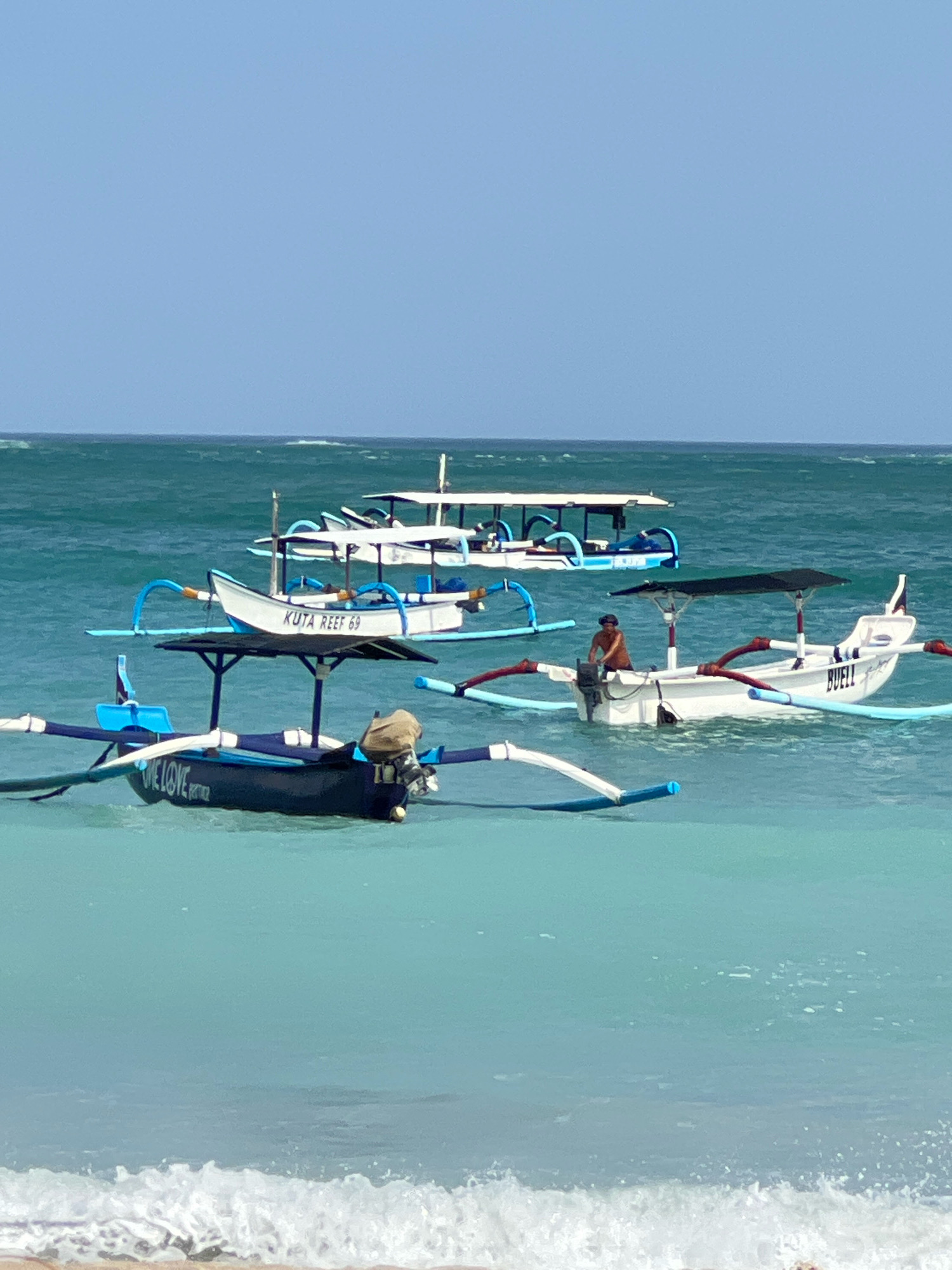 In Kuta, Indonesia, this vertical, medium shot captures several traditional Balinese outrigger boats floating on turquoise waters under a clear, pale blue sky. The foreground features a gently breaking wave washing onto a sandy shore. In the middle ground, four boats are visible. The closest boat, predominantly dark blue with white trim, has "ONE LOVE" inscribed on its side and a beige bag with an outboard motor attached. Behind it, a white boat with blue trim, labeled "KUTA REEF 69," is positioned. To the right, another white outrigger boat with a dark canopy has a person in a brown shirt operating its outboard motor and the name "BUELL" visible on its side. Further in the background, another boat with a white canopy is partially obscured. The boats are characteristic of those used for local transportation and tourism in the region.