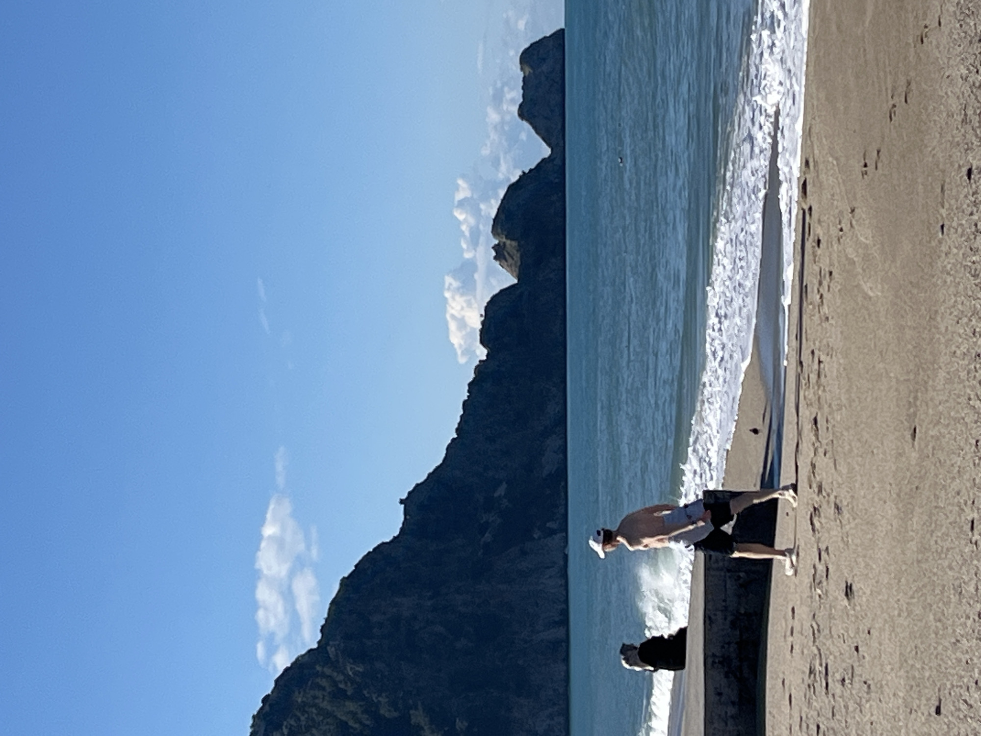 The image depicts a coastal scene in Alanya, Turkey. A person wearing shorts and a hat is standing on a sandy beach. Waves are crashing on the shore next to them. In the background, a large, dark, rocky landform rises from the sea. The sky is blue with some clouds.