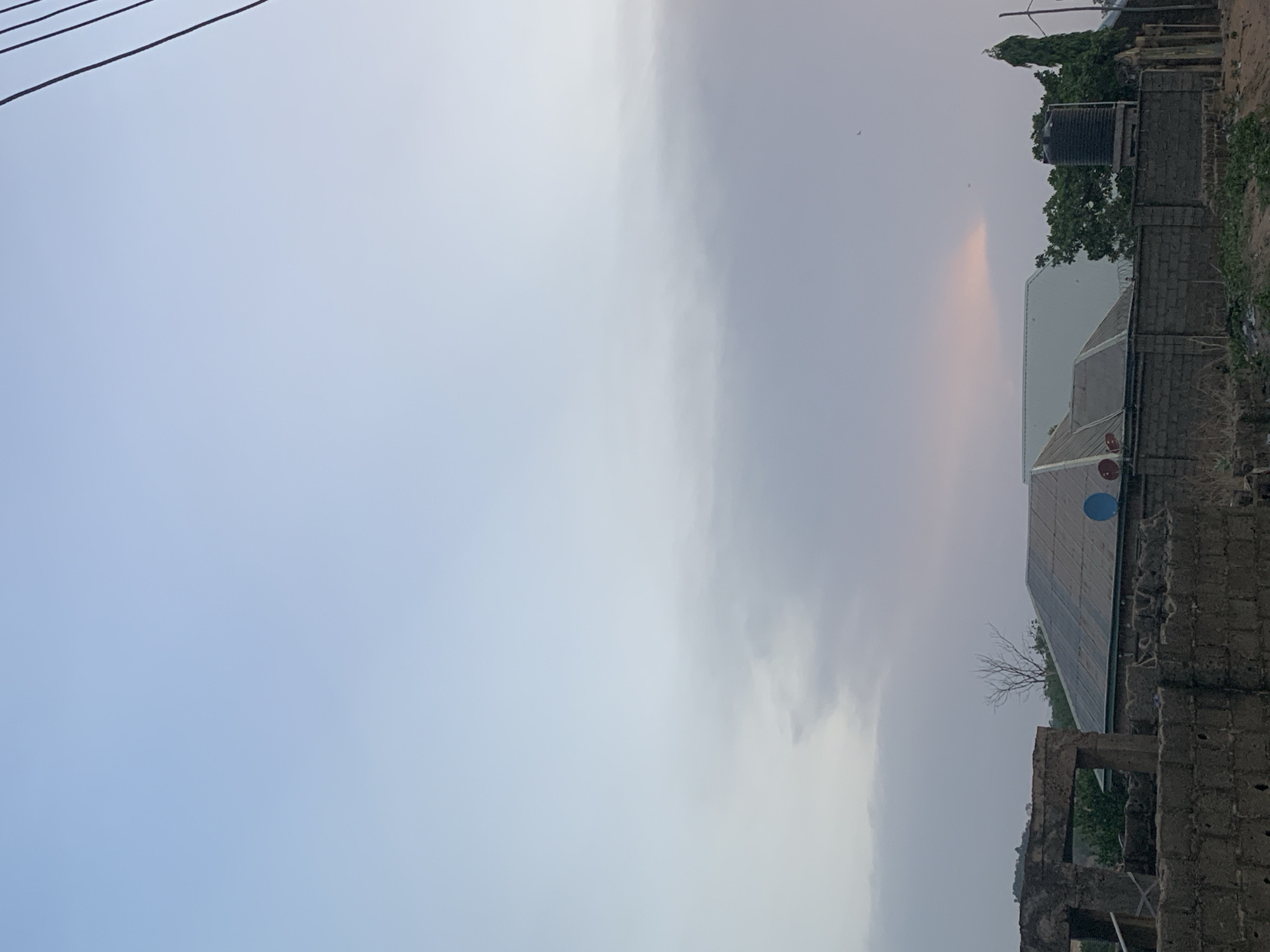 The image shows a street scene in Lokoja, Nigeria. The sky is overcast with light, diffused sunlight. In the foreground on the right, there are structures including brick buildings and a zinc roofed building with a blue satellite dish. Adjacent to the building there is foliage, including a tree. A black water tank is visible on the roof. Power lines are visible in the upper left.