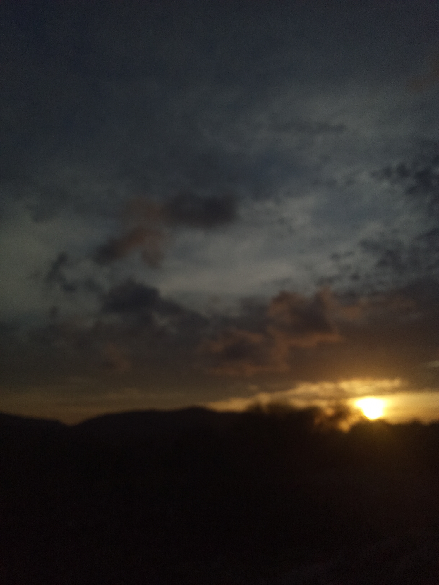 The image shows a sunset in Altagracia, Venezuela. The sky is filled with dark, layered clouds. The sun is setting on the horizon, obscured by trees and mountains. The foreground is dark and indistinct.