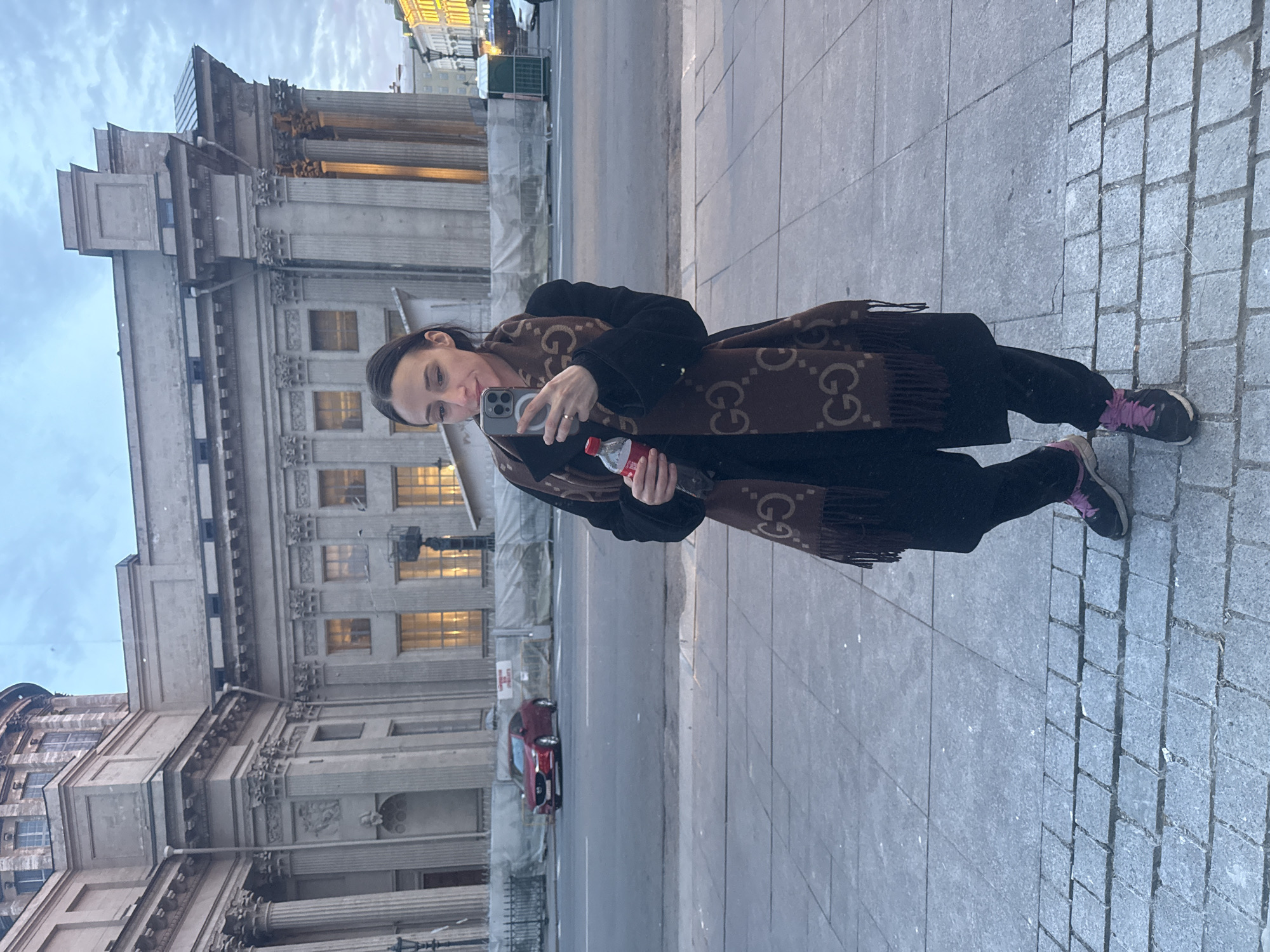 A woman stands on a cobblestone sidewalk, angled towards the camera, holding a phone in her right hand and a bottle of Coca-Cola in her left. She wears a dark coat, dark pants, and sneakers with pink laces. A brown scarf with a "G" pattern is wrapped around her neck. Behind her is a multi-story building with classical architectural features, including columns and ornate detailing. A red car is visible in the street in front of the building. The sky is overcast. The location is Saint Petersburg, Russia, specifically in front of the General Staff Building on Palace Square.