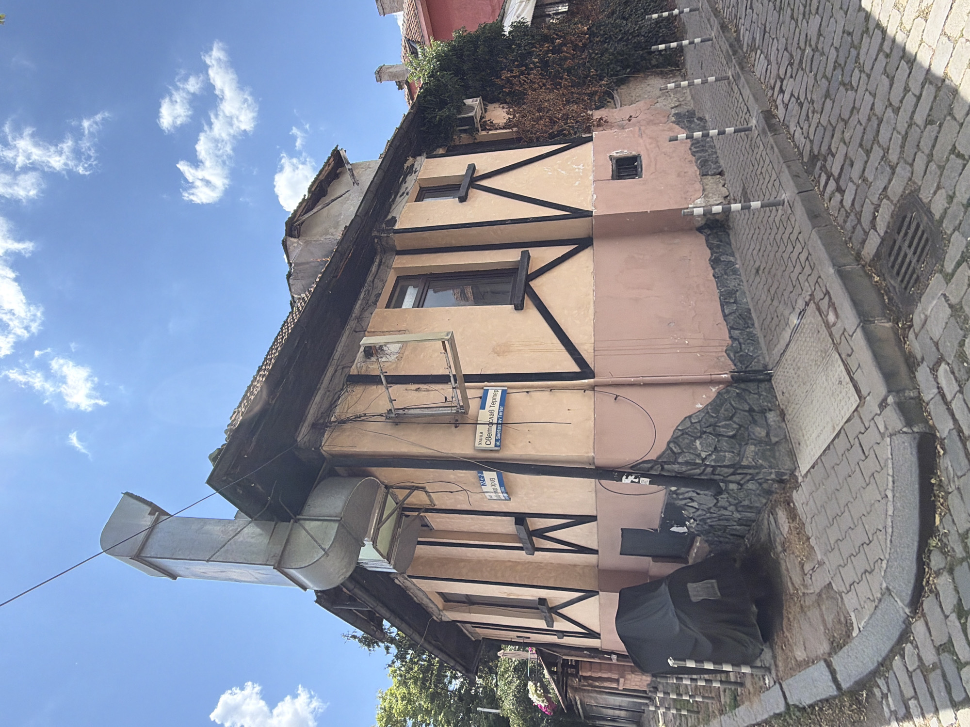 This image captures a street view in Sofia, Bulgaria, specifically on Svetoslav Terter Street. The focal point is a two-story building with a distinctive architectural style featuring a pale pink facade and dark wooden beams forming a decorative pattern on the upper story. A large, angular metal ventilation duct extends from the side of the building upwards, dominating the upper left portion of the frame. The building has several windows, some with dark wooden frames. The lower section of the building's exterior is clad in dark, irregular stone.
The street is paved with cobblestones, and a curved curb separates it from the sidewalk. Several striped bollards are positioned along the sidewalk. In the foreground, to the left, a dark tarp covers an unseen object, possibly a vehicle or equipment. The sky is bright blue with scattered white clouds. To the right of the building, dense green foliage and some dry, brown vegetation are visible, along with an air conditioning unit mounted on the wall. A red building is partially visible further down the street. The overall scene suggests a historical urban setting.