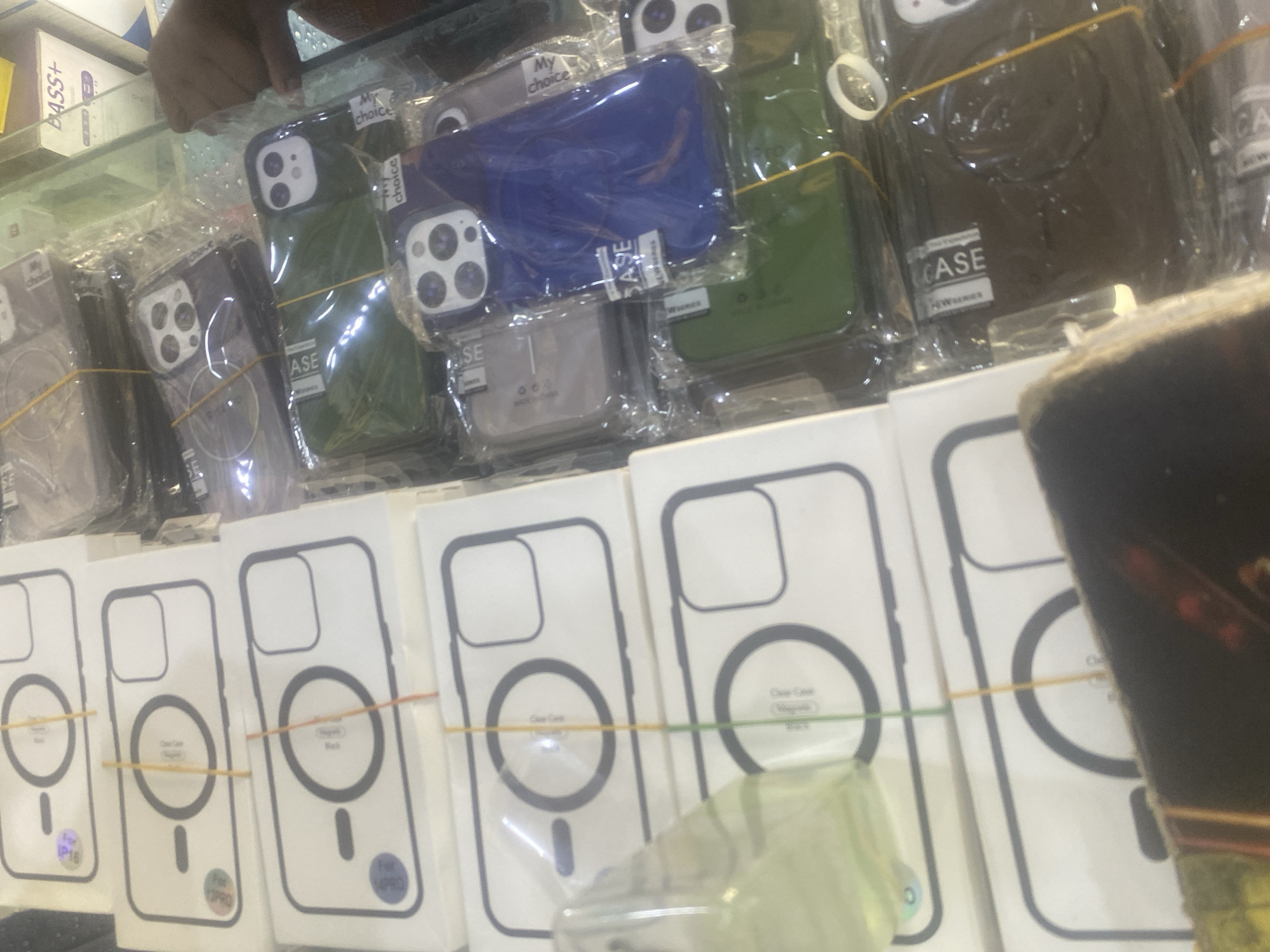 The image displays a close-up view of a retail display, likely in a phone accessory shop in Jalingo, Nigeria. The primary subjects are numerous mobile phone cases, predominantly for iPhones, arranged in plastic packaging. Some cases are visible outside their packaging, showcasing different colors like green, blue, and black, and designs, some featuring multiple camera cutouts indicative of higher-end phone models.
Several white boxes are also prominent, displaying a silhouette of a phone case with a circular magnetic charging area. These boxes are banded together with colorful rubber bands. Visible text on the packaging includes "My choice," "CASE," "ASE," "For IP16," and "NWAERIES," suggesting branding and product information. In the background, a person's hand and arm are partially visible, suggesting they might be a shopkeeper or customer. The lighting appears to be indoor, typical of a shop environment. The overall scene conveys a commercial setting where mobile phone accessories are being sold.