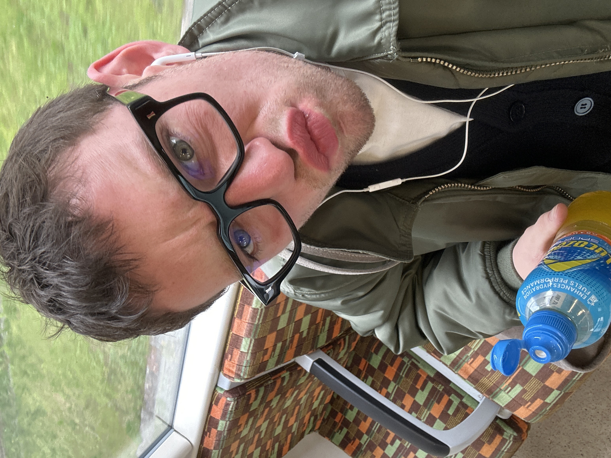 A man in a green jacket and black framed glasses is on a train in London, UK. He has an earbud in his ear and is holding an open bottle of Lucozade. The train seat has a patterned cushion. Outside the train window is a blurred view of greenery.