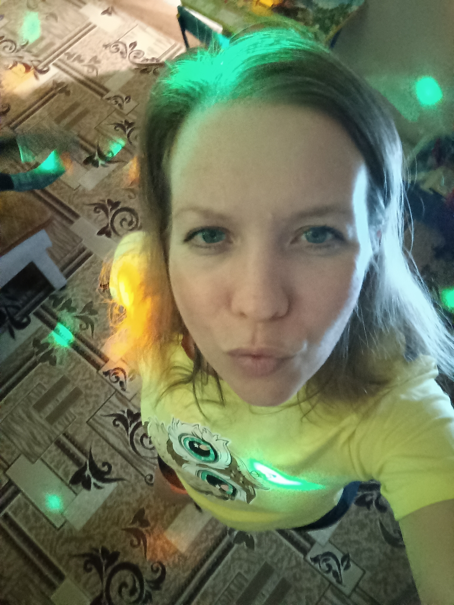 A woman is taking a selfie from a high angle, looking down at the camera. She has fair skin, blue eyes, and light brown hair. She is wearing a yellow t-shirt with a cartoon owl design on the front. The background is a room with a patterned floor. There is a small fireplace or heater visible in the lower left corner. There are several green light effects scattered throughout the image, likely from a light source. The location is Chaikovsky, Russia. There are no discernible actions or interactions besides the woman taking a selfie.