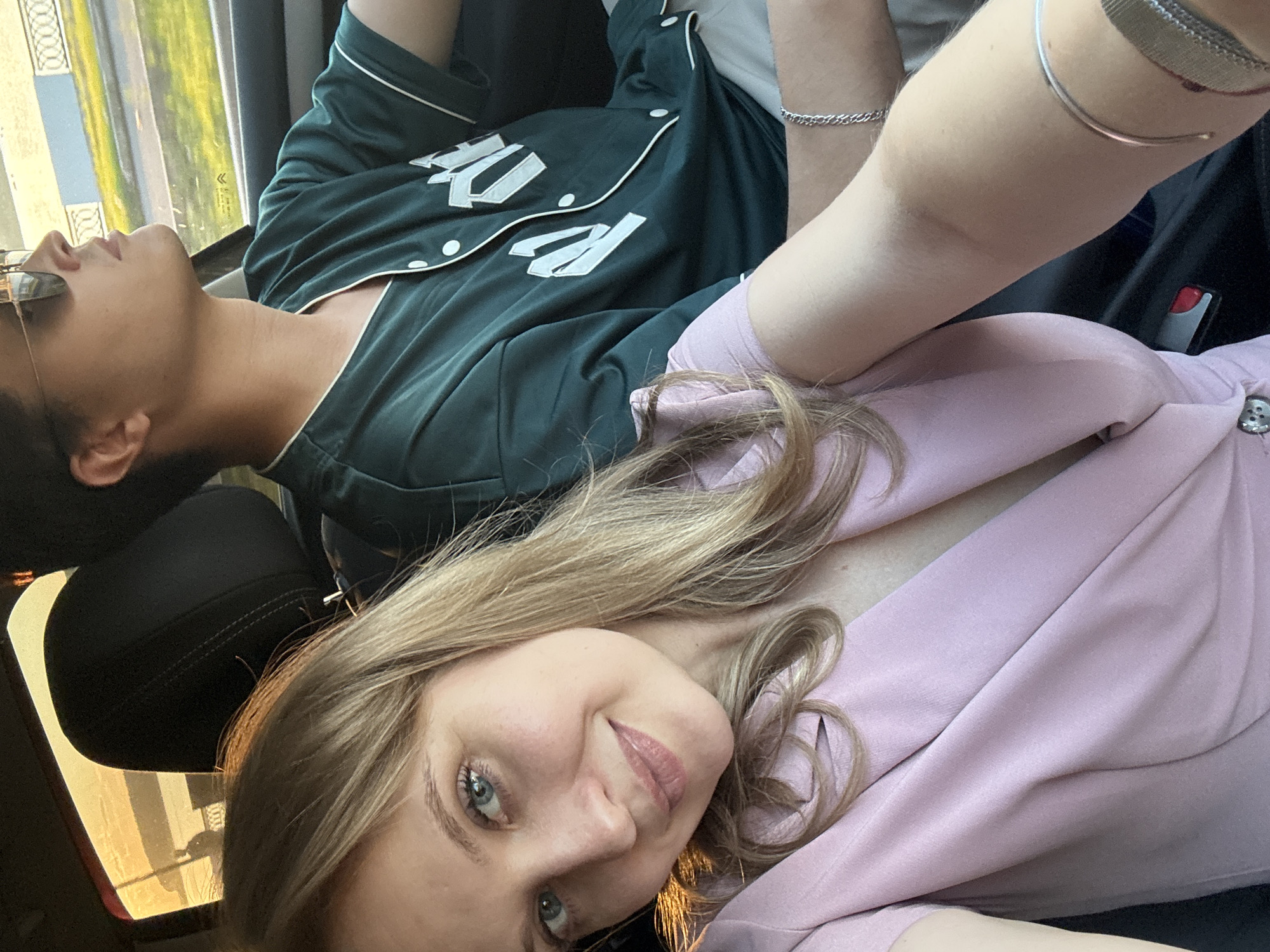 The image depicts two individuals inside a moving vehicle, identified as a Citroën, in an urban setting within Izhevsk, Russia, under warm natural light, likely late afternoon.
In the foreground, a woman with blonde hair, blue eyes, and light makeup smiles directly at the camera, holding a device to capture a selfie. She wears a light pink V-neck top or dress with a visible silver button closure near her left arm. Her left forearm displays a faint scar and is adorned with a simple silver bangle bracelet and a watch with a silver mesh band.
Behind her, to the right, a man with dark hair is partially visible in profile, wearing reflective sunglasses and a dark green, short-sleeved, button-up shirt with white piping and white letters or numbers (partially visible, possibly "M" and "P" or "R"). A silver chain-link bracelet is on his left wrist. He is oriented forward, looking ahead through the windshield, presumably driving. The car's interior features dark headrests and trim.
Through the car windows, an urban street environment is visible. The man's side reveals an asphalt road, a gray curb, a grassy median with some yellowed patches, and a gray-blue metal fence with circular top elements. In the distance, a light-colored building and some trees are discernible. The overall lighting suggests a clear day with the sun low in the sky.