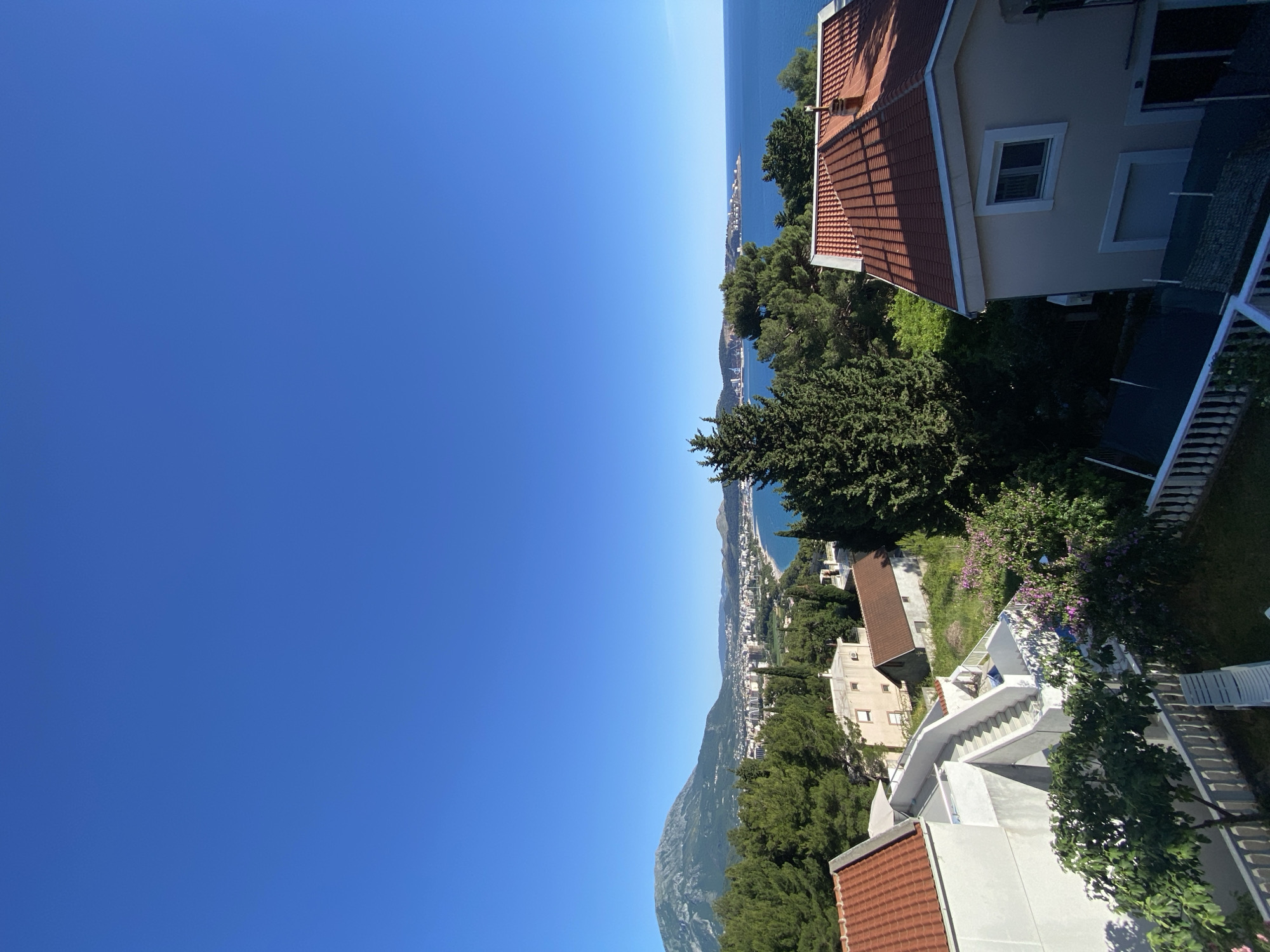 The image shows a view from a high vantage point in Šušanj, Montenegro. The landscape includes residential buildings with red-tiled roofs nestled amongst green trees. In the distance, the Adriatic Sea is visible. The sky is clear and blue. A city skyline can be seen along the coast in the middle distance. A mountain is visible near the bottom left. Some stairs can be seen leading down a slope. The photograph appears to have been taken during the day in sunny weather.