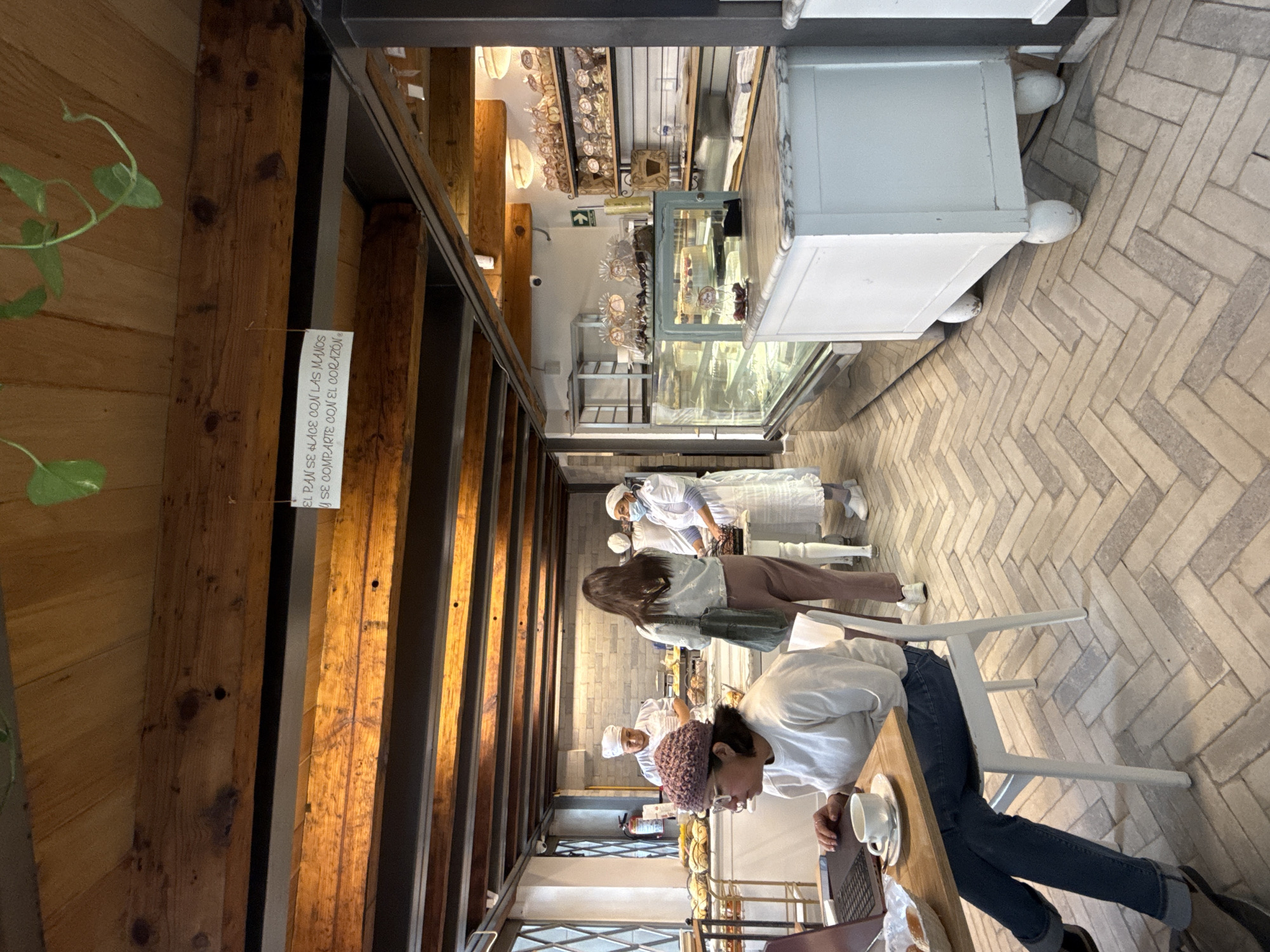 A cafe or bakery in Mexico City, Mexico. The scene is an interior view, with light brown wooden beams and a wooden wall on the left. A white sign is attached to the beam.
The right side of the image shows a white counter with a glass display case. The floor has a brick pattern. Several people wearing white uniforms and hats are behind the counter, potentially staff. A woman in a gray sweater and brown pants is standing next to the counter.
In the foreground, a woman in a white sweater and jeans is sitting at a table working on a laptop, with a cup and saucer.
The overall environment suggests a modern, upscale bakery or cafe setting.