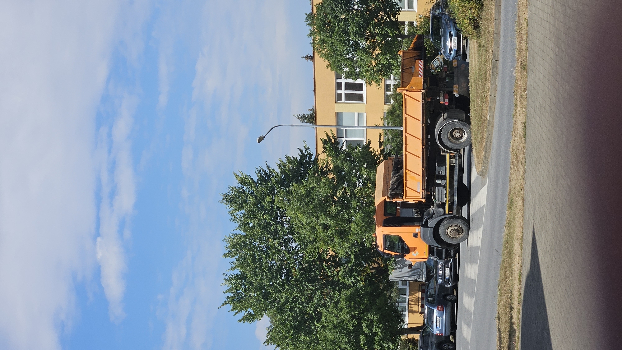 An orange dump truck is parked on a paved road in front of a yellow building. The truck has a large, open bed with a tarp covering part of its contents. Several cars are parked on the side of the road, and trees with green foliage line the area. A lamppost stands near the building. The sky is blue with scattered clouds. The scene appears to be on a street in the city of Prešov, Slovakia.