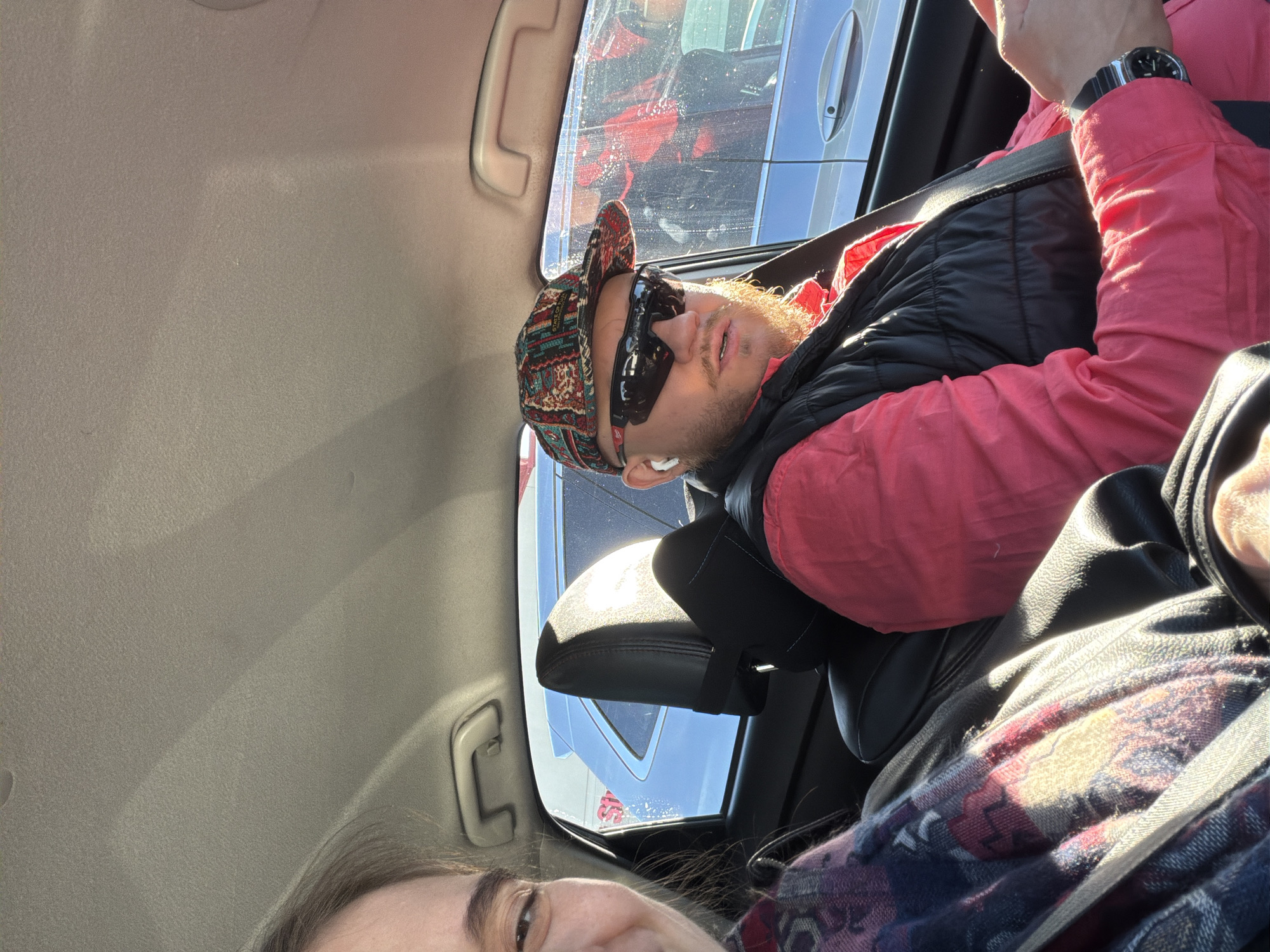 The image shows two people inside a car. The person in the front passenger seat is a man with a beard, wearing a patterned bandana, sunglasses, a red shirt, a black vest, and a watch. He has an earpiece in his ear and is wearing a seatbelt. The driver is only partially visible, showing a portion of their face and a patterned scarf. The view out the car window reveals other vehicles, suggesting a parking lot or traffic situation. The location is in Niles, United States of America, as inferred by the image metadata.