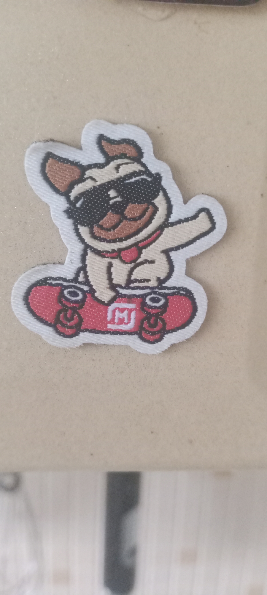 A fabric patch depicts a cartoon pug wearing sunglasses and a red collar, riding a red skateboard. The pug has its tongue sticking out and its right arm raised. The skateboard has four wheels and a white emblem with the letter "M" inside a square. The patch has a white border and is placed on a light brown, textured surface. The background is out of focus, showing vertical lines. The location in Kudymkar, Russia, is not discernible from the image.