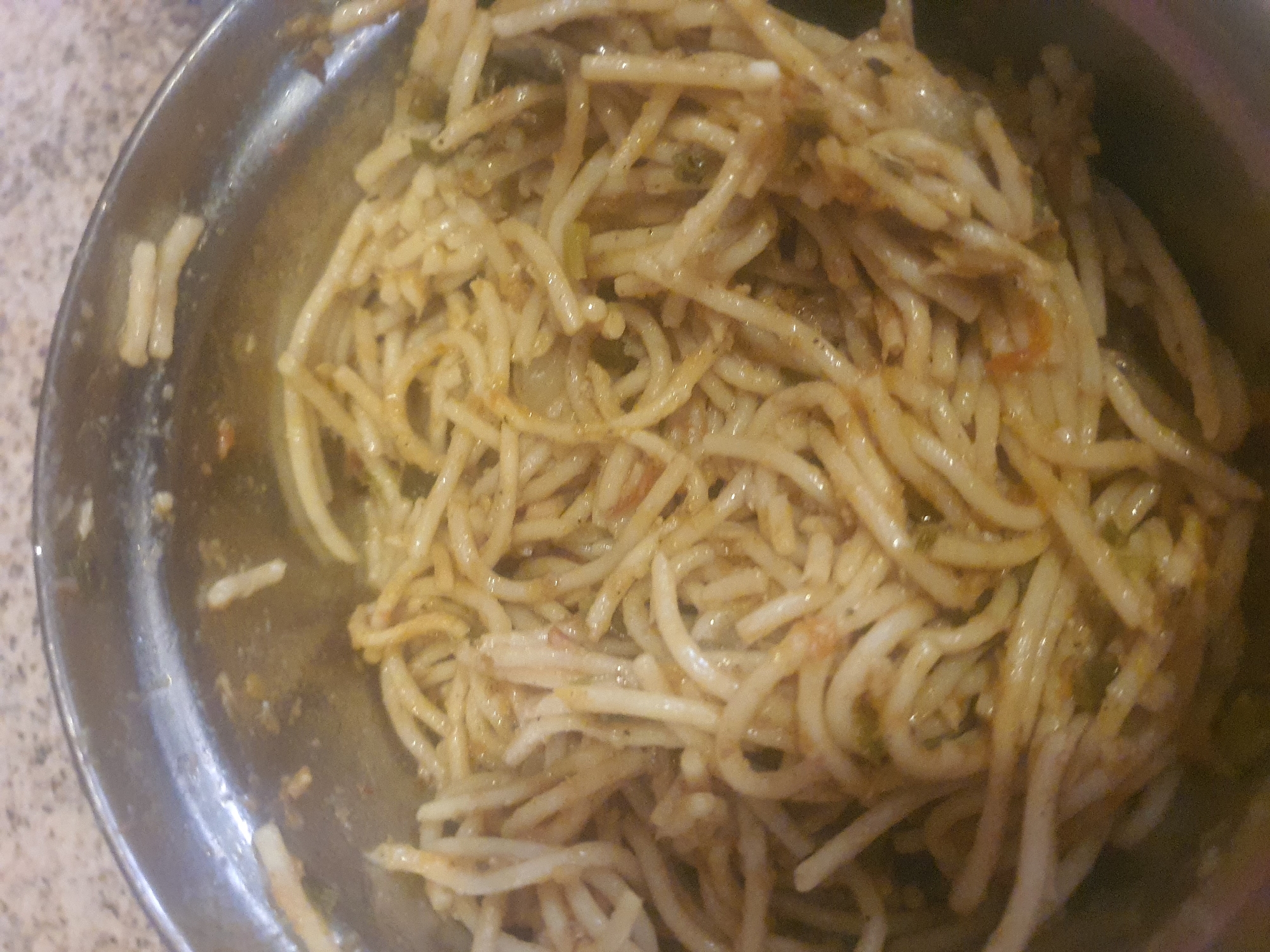 A close-up shot shows a metal bowl filled with cooked noodles. The noodles are thin and appear to be coated in a sauce, with visible pieces of what might be vegetables mixed in. The bowl itself is a dull silver metal and is positioned on a speckled surface, possibly a countertop. The lighting in the image suggests it was taken indoors, with no indication of the time of day or weather conditions. There is no visible text in the image. The image focuses entirely on the food, with no people or other objects in the frame. The setting appears to be a domestic or casual dining environment in Jos, Nigeria, given the provided location context. The noodles are the sole subject of the image.