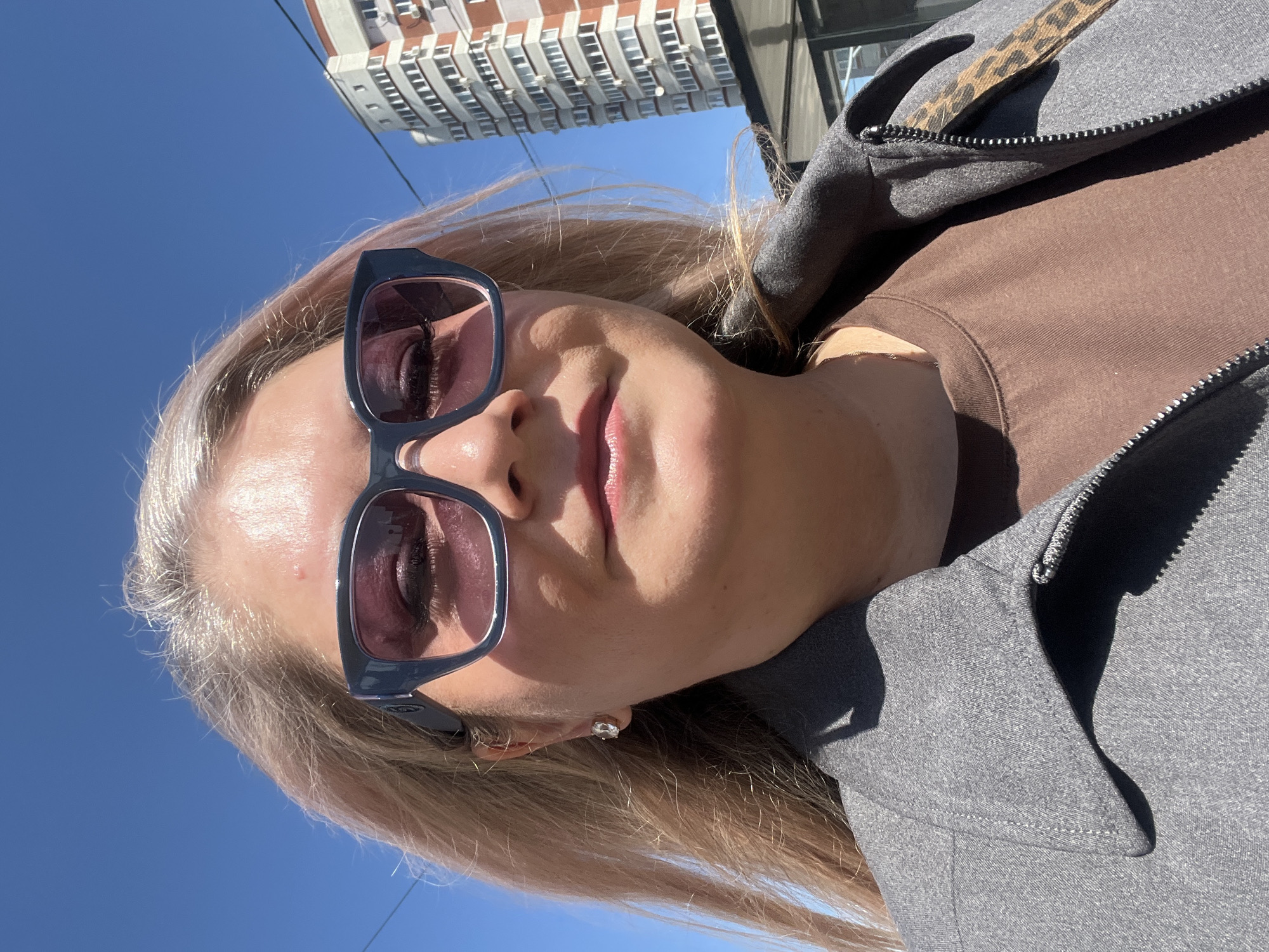 The image is a close-up selfie of a woman wearing sunglasses. She has light blonde hair and is wearing a brown shirt with a gray jacket. In the background, there is a clear blue sky and a tall residential building with multiple windows. Based on the architectural style of the building, the location may be Izhevsk, Russia.