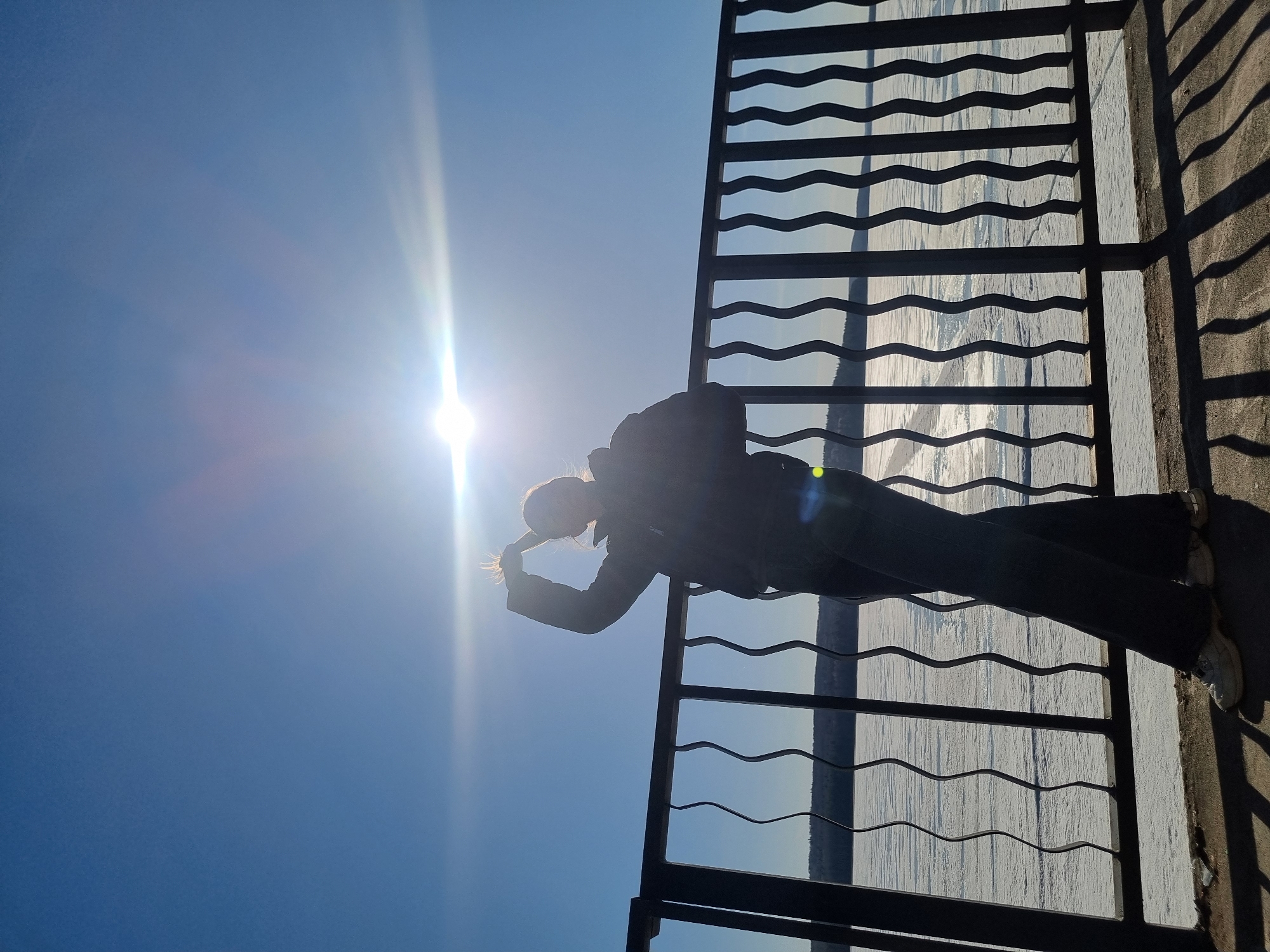 A person stands in profile, silhouetted against a brilliant sun in a clear blue sky. The image is composed with a strong backlight, causing the subject and a metal railing to appear as dark outlines. The person, who appears to be female, is wearing dark clothing and light-colored shoes, with one hand raised towards her head, possibly adjusting her light-colored hair or shielding her eyes.
She is positioned next to a dark metal railing that features distinctive horizontal bars with a wavy, undulating pattern. This railing runs across the right side of the frame, casting long, patterned shadows onto the concrete or paved ground where the person stands. Beyond the railing, a vast expanse of water, likely a river or reservoir, is visible. This body of water appears to be covered in ice or snow, creating a textured, reflective surface under the bright light.
The sun is a powerful light source in the upper left, generating significant lens flare and long, distinct shadows, indicating either early morning or late afternoon. The sky is a uniform, clear blue, suggesting fair weather, though the icy water implies a cold environment. The scene depicts a serene moment at what appears to be an observation deck or promenade overlooking a frozen landscape, fitting for a location such as Chaikovsky, Russia. No visible text is present in the image.