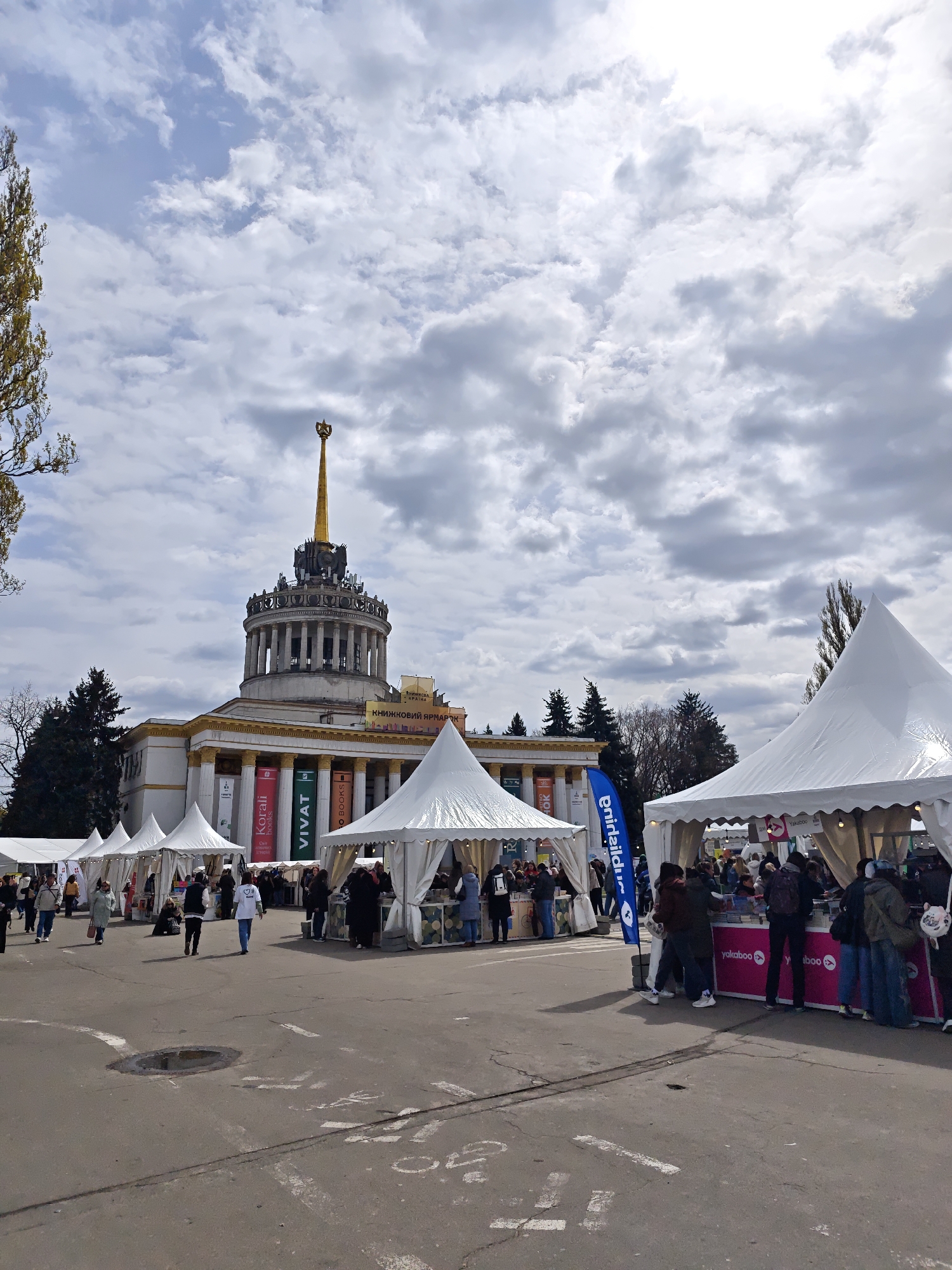 The image captures an outdoor book fair in Kyiv, Ukraine, set against the backdrop of a grand, classical-style building under a cloudy sky. The central building is a prominent architectural feature, characterized by a large dome topped with a distinctive golden spire and surrounded by columns.
Numerous white, peaked vendor tents are arranged in rows on a paved plaza in front of the building. Many people are visible throughout the scene, casually dressed and engaged in the activities of the fair, either walking between tents, browsing merchandise, or congregating around specific stalls.
Banners draped from the building's facade clearly indicate the event's nature with text such as "КНИЖКОВА КРАЇНА" (Book Country) and "КНИЖКОВИЙ ЯРМАРОК" (Book Fair). Other visible vendor names on banners include "Korali books," "VIVAT," and "O BOOKS." Near one of the tents, a display shows "yakaboo," a Ukrainian online bookstore. A blue vertical flag with partially visible text is also present.
The sky is heavily overcast with a mix of white and grey clouds, suggesting a mild, daytime atmosphere. Sparse, yellowed leaves on trees to the left hint at either autumn or early spring. The ground is paved, showing some road markings and a small puddle. The overall impression is one of a bustling public event.