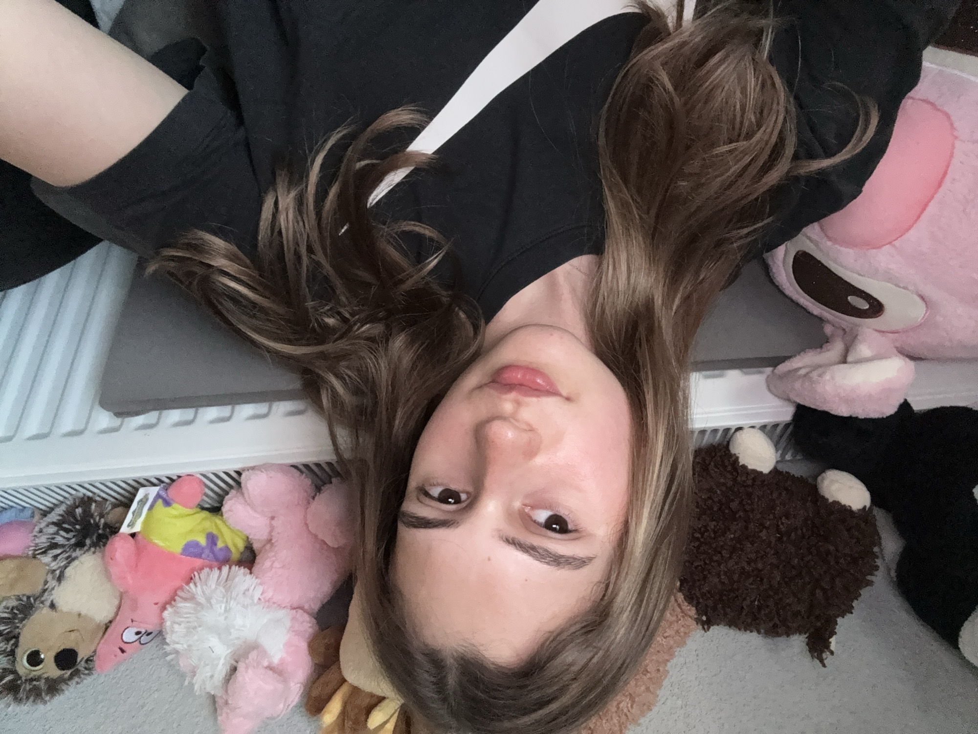 A young woman is lying on her back, surrounded by stuffed animals. She is wearing a black Nike T-shirt. Her hair is brown and parted in the middle. The background shows a radiator and carpet. There is no indication of this image being taken in Panagyurishte, Bulgaria.