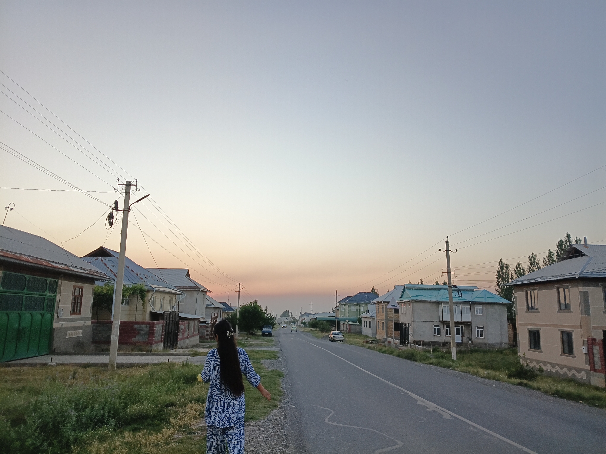 In Uchkun, Kyrgyzstan, at dusk, a woman in a blue and white patterned outfit walks along a paved road lined with houses. The sky is a pale gradient of blue and orange, indicating the setting sun. Utility poles and wires run alongside the road. Several cars are visible on the road, and the houses on either side of the street vary in architectural style and color. Green vegetation grows along the roadside and in front of some of the houses.