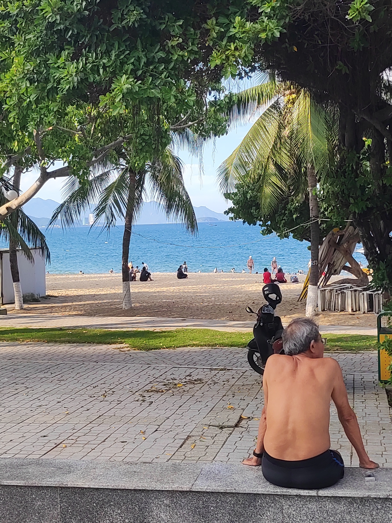 The image depicts a scene on a beach in Nha Trang Ward, Vietnam. In the foreground, an older man with graying hair is sitting on a concrete ledge, facing away from the camera and looking towards the ocean. He is shirtless, wearing black shorts, and appears to be relaxed. To his left, a black scooter is parked on a paved walkway.
Beyond the walkway is a grassy area, and then a sandy beach with several people scattered about. Some individuals are sitting or standing near the water's edge, while others are further up on the sand. The ocean is a vibrant blue, and in the distance, mountains and a few buildings are visible on the horizon. Palm trees and lush green foliage frame the scene, casting shadows on the beach and walkway. The overall impression is one of a peaceful, sunny day at a tropical beach.