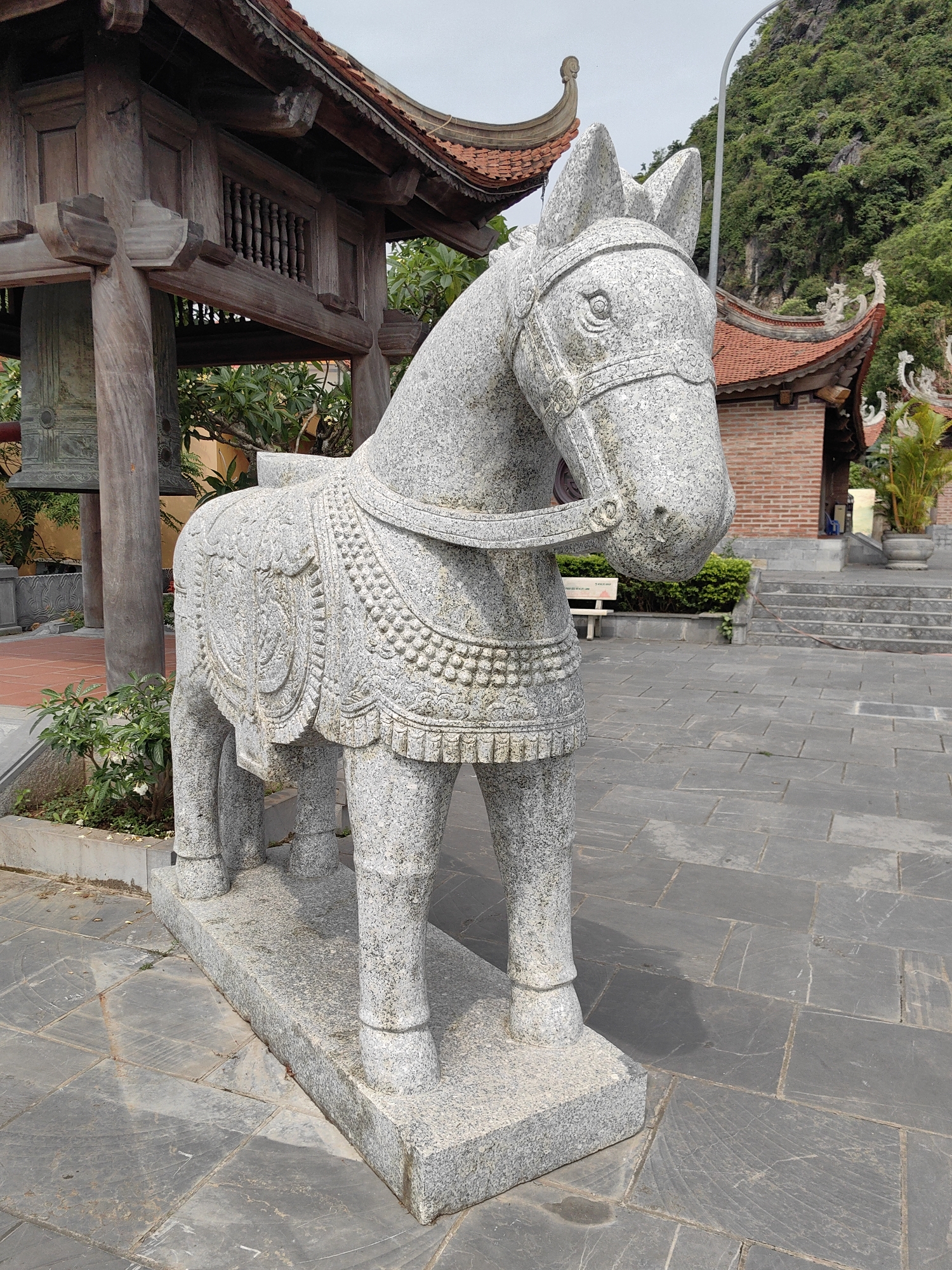 A stone statue of a horse, ornamented with carvings resembling saddle blankets and bridle, stands on a stone pedestal in what appears to be a courtyard. The courtyard is paved with large, flat gray stones. In the background are traditional Vietnamese architectural elements, including a wooden structure with a tiered roof, a brick building with an orange tile roof, steps leading up to the building, and decorative sculptures along the roofline. Lush green foliage and the base of a mountain covered in trees are visible in the distance. The scene is located in Hong Gai Ward, Vietnam. The weather appears overcast or hazy.