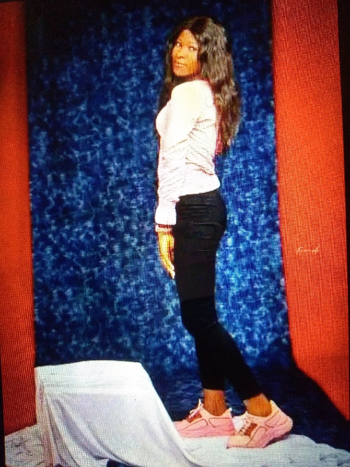 A woman stands in a studio. She wears a striped shirt, dark pants, and pink sneakers. Her dark hair falls over her shoulders. She gazes over her shoulder at the viewer. Behind her is a blue, textured backdrop and a white-draped object. Red vertical bars flank each side of the backdrop.