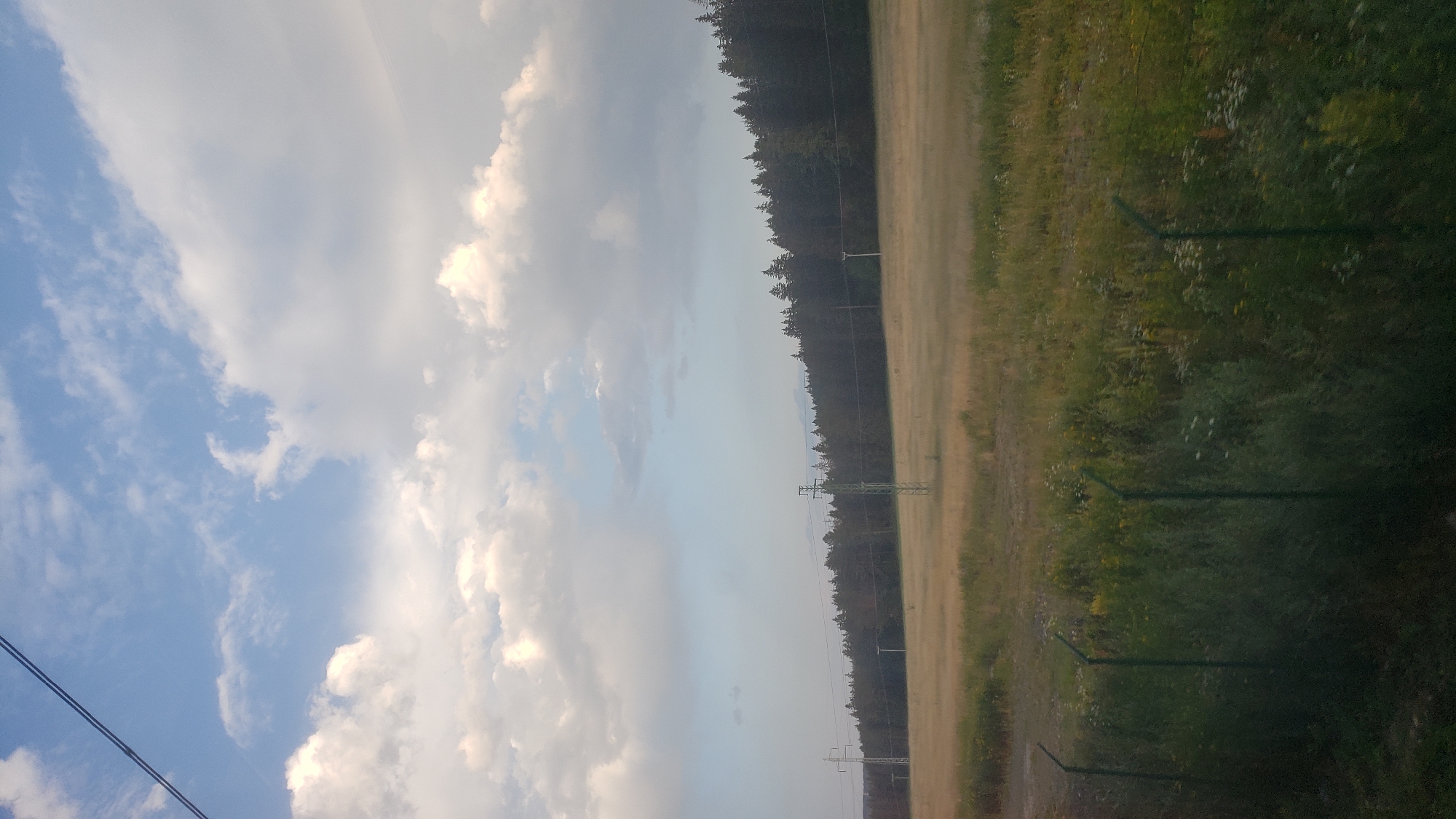 The image shows a wide, open field in Lučivná, Slovakia, with a dark forest bordering the far side. A chain-link fence runs along the bottom of the frame, separating the viewer from the field. In the distance, power lines and electrical towers are visible, crossing the field and disappearing into the trees. The sky above is partly cloudy, with a mix of white and gray clouds against a light blue background. The overall impression is of a rural landscape under a dynamic sky.