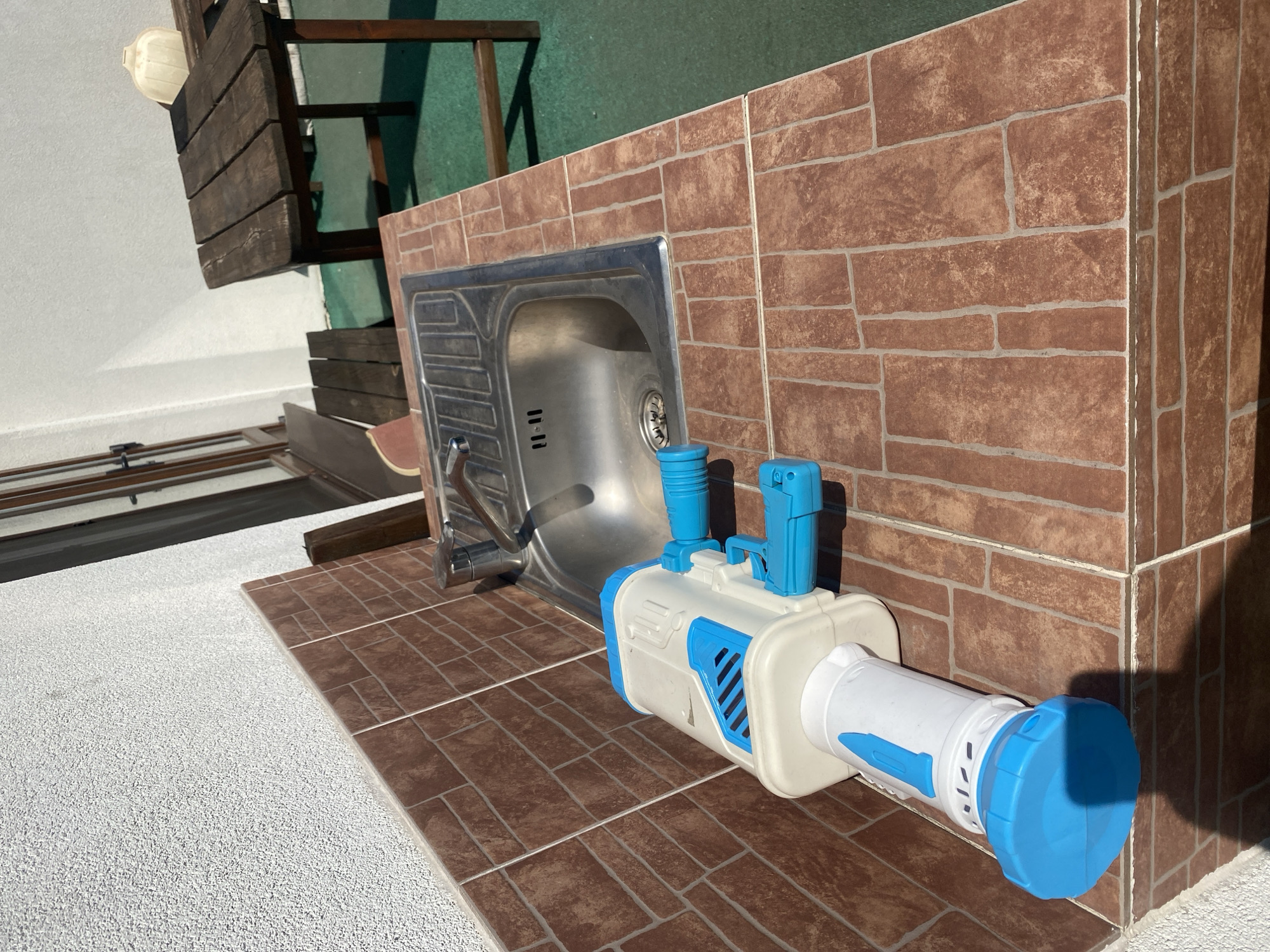 A close-up, high-angle shot shows a blue and white toy water gun resting on a tiled countertop next to a stainless steel sink. The sink has a faucet and what appears to be the numbers "11-11" etched into its surface. The countertop is made of reddish-brown tiles laid in a brick pattern. Behind the sink, a portion of a wooden table with a green surface beneath it is visible. The background includes a white textured wall and a window with wooden frames. The scene appears to be an outdoor kitchen or patio area, potentially located in Chișinău, Moldova.