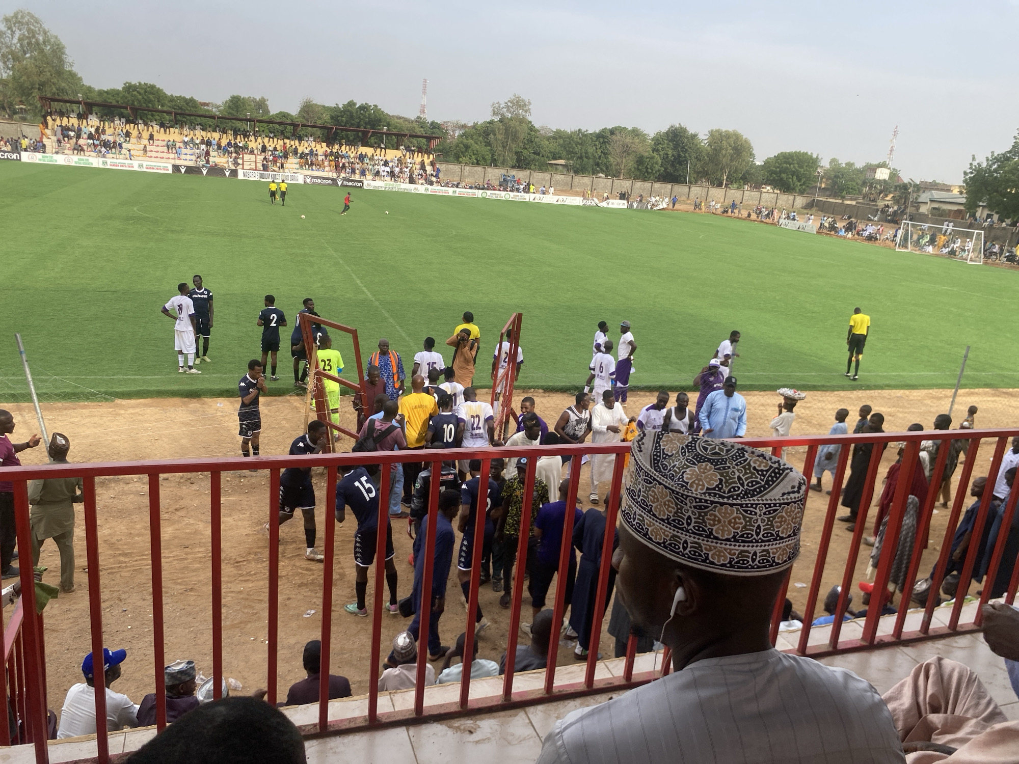 The image shows a soccer match in progress at a stadium. The stands are partially filled with spectators. Players in various team uniforms are present both on the field and near the barrier in front of the seating area where the photo was taken. A man wearing a traditional patterned cap is in the foreground. Based on the stadium signage, the location is Nasarawa Stadium, Tudun Wada, Zaria, Nigeria.