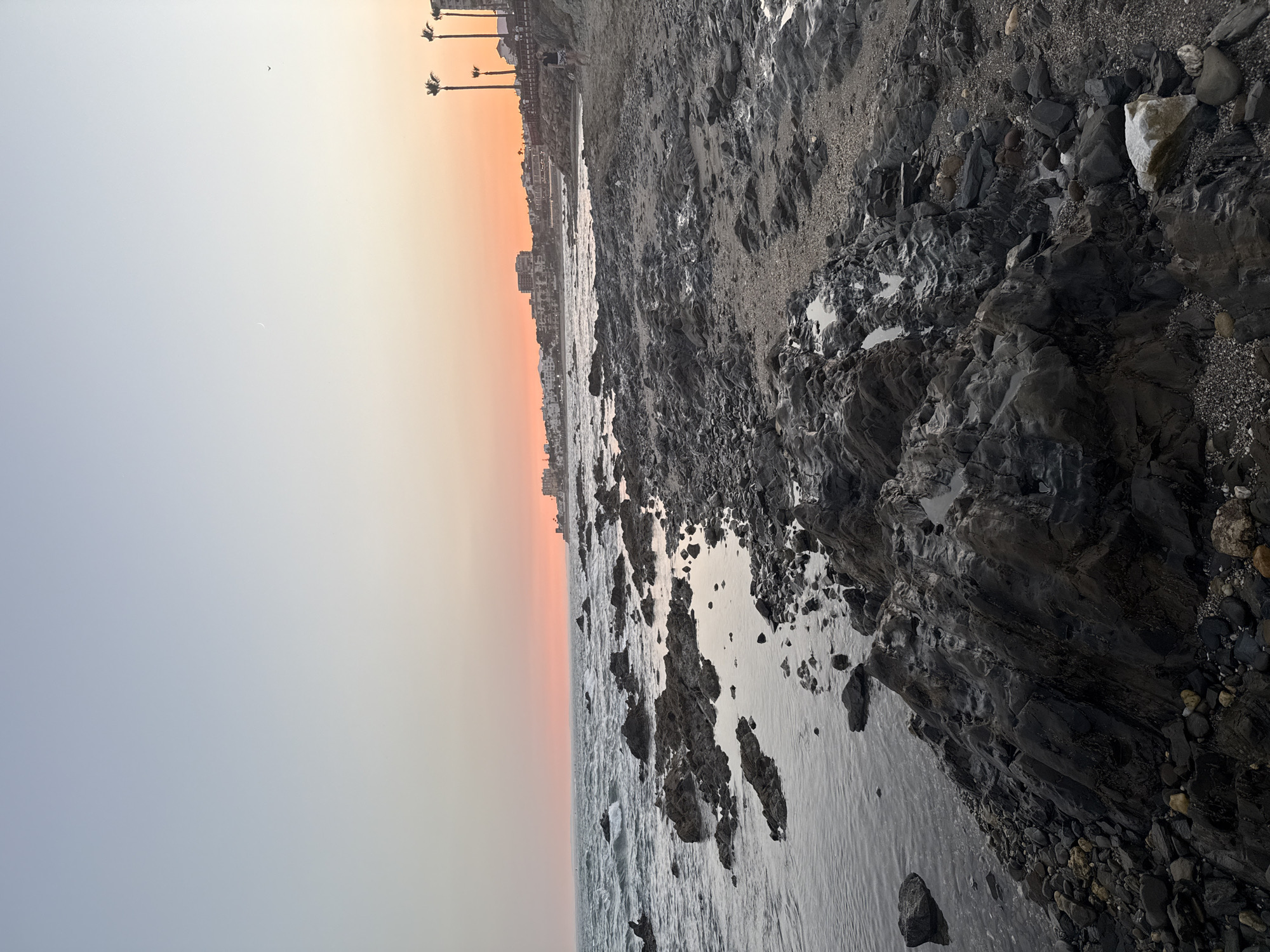 The image depicts a rocky beach with the ocean on the left and a sandy area with scattered rocks on the right. The sky exhibits a gradient of colors, transitioning from a pale blue at the top to a soft orange and pink near the horizon, indicating either sunrise or sunset. A crescent moon is visible in the upper left portion of the sky. In the distance, a coastal town with buildings and palm trees lines the shore. A few people are visible walking along the boardwalk and beach area. The foreground is dominated by dark, wet rocks and small tide pools reflecting the sky. The overall scene is a natural coastal landscape with a developed town in the background, suggesting it is a populated area of Mijas, Spain.