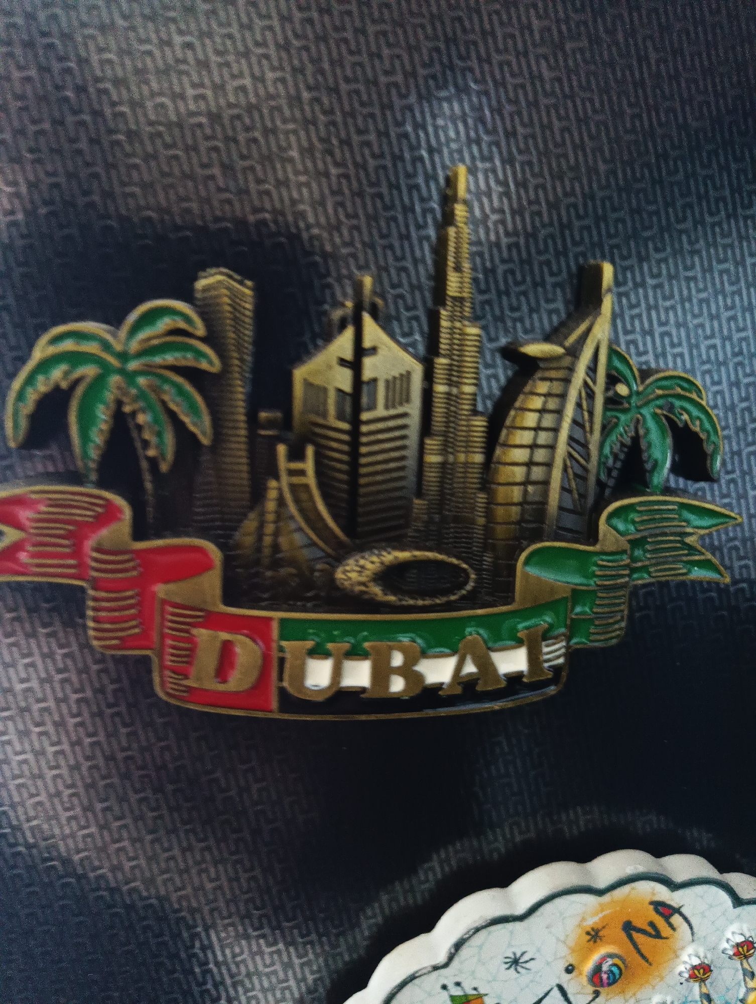 The image displays a Dubai-themed souvenir magnet, featuring a miniature representation of the city's iconic skyline. Prominent structures such as the Burj Khalifa and the Burj Al Arab are visible, alongside palm trees, all rendered in gold and green enamel. A banner across the bottom of the magnet reads "DUBAI" in gold lettering against a red and green background. The magnet is placed on a textured, dark blue surface, possibly a fabric or patterned wallpaper. There is no indication or context suggesting the city of Karnobat, Bulgaria, in this image.