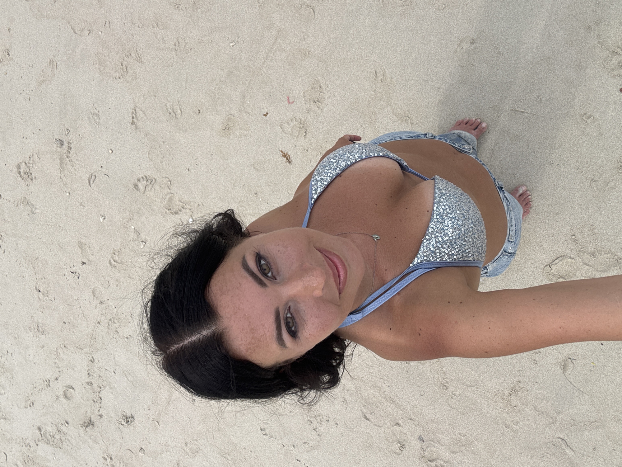 A woman stands on a sand beach. She wears a silver bikini top and light-wash denim shorts. The beach sand is beige with visible footprints. Her hair is dark, and she gazes upward into the camera. This image was likely taken in Ban Haad Rin, Thailand.