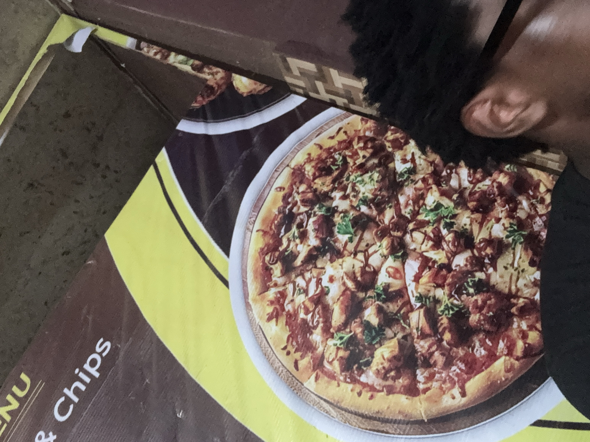 The image depicts a close-up of a takeaway menu for a food establishment, likely a restaurant or fast food outlet, located in Eziobodo, Nigeria. The menu features an image of a pizza with a brown and white topping, cut into slices. Partial text visible on the menu includes "& Chips". A person's ear and some hair are visible in the upper right portion of the image, partially obscuring the pizza. A cardboard box is also partially visible in the top left corner of the frame.