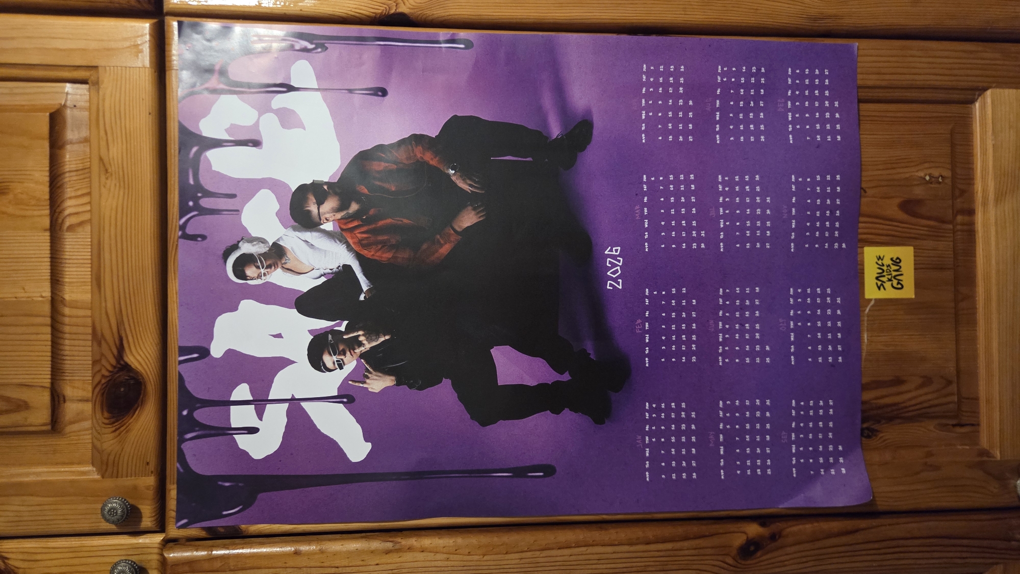 A poster is hanging on a wooden cabinet. The poster features three people in front of a purple background with white text and dripping purple graphics. The people are dressed in streetwear. The bottom portion of the poster contains a calendar for the year 2026, with each month displayed in a grid format. There is a yellow sticky note with the text "SAUCE KIDS GANG" adhered to the bottom right corner of the poster. The wooden cabinet has two decorative knobs on the top panel.