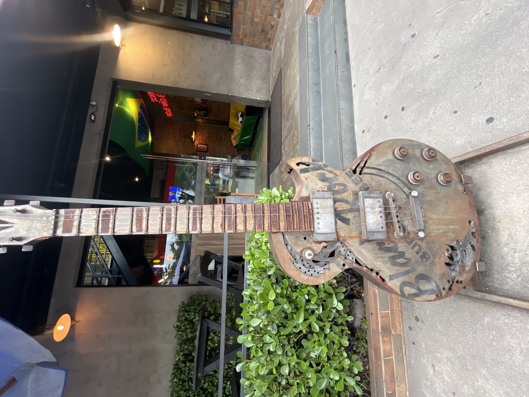 A full-frame shot shows a rusted, metal guitar sculpture positioned in front of a building in an unknown city. The guitar is positioned vertically, with the neck of the guitar extending to the left and the body of the guitar to the right. The guitar appears to be crafted from metal and has a heavily rusted and weathered appearance. The word "Blues" is prominently displayed on the guitar body. The sculpture is surrounded by green plants. Behind the guitar, the entrance of a building is visible, with neon signs and interior decor suggesting a bar or restaurant. The building has brick and concrete elements. To the right of the guitar, there is a sidewalk or pavement area.