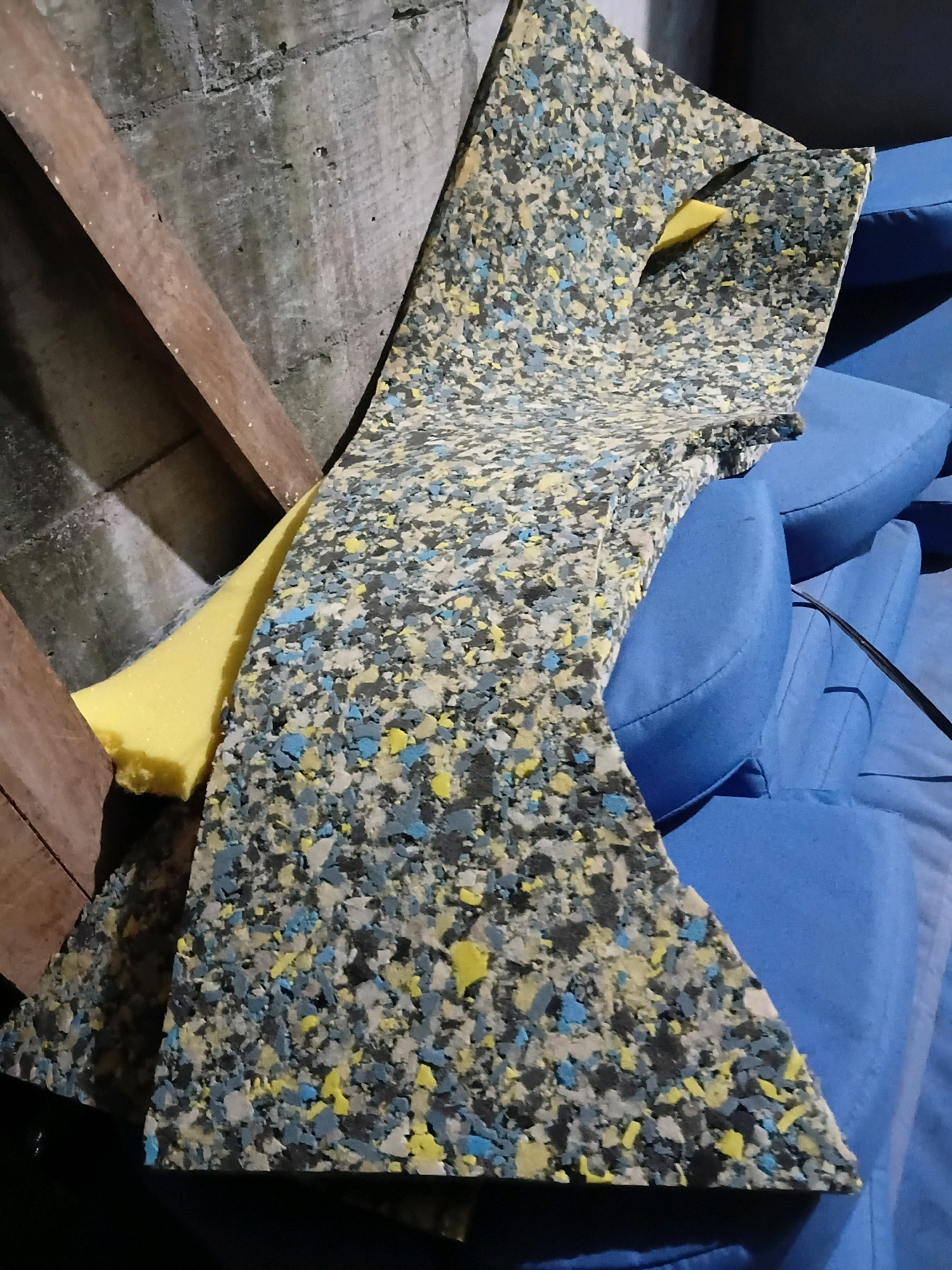 The image depicts a close-up view of several pieces of foam padding, some of which are layered and cut into irregular shapes. The foam is a composite material, appearing as a mottled pattern of black, grey, yellow, and blue fragments. A bright yellow piece of foam is partially visible underneath a larger, more complexly shaped piece of the same composite foam. This composite foam is draped over what appears to be a stack of blue upholstered cushions, which are slightly out of focus and form the background of the image. The overall scene suggests a storage or workshop area. To the left, a rough concrete wall is visible, with a wooden plank leaning against it. The lighting is dim, casting shadows and highlighting the textures of the materials. There is no indication of the specific location within the city of Cikal, Indonesia, nor any people or actions taking place.