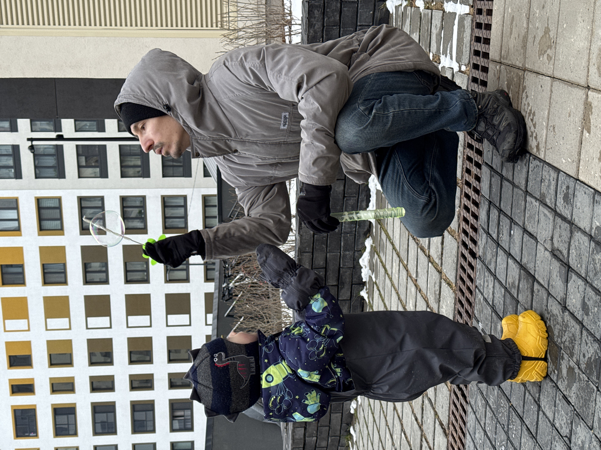 A man and a child are outside in what appears to be a courtyard or walkway in a city setting, possibly Izhevsk, Russia, given the urban architectural style. The man is in a crouching position, wearing a gray hooded jacket, black gloves, and jeans, and he is blowing a bubble using a green bubble wand. A small, clear bubble is visible near the wand. The child is standing upright near the man, wearing a dark jacket with colorful prints of rockets, a gray hat with decorative details, black pants, and bright yellow boots. The background includes a modern building with a grid of windows, as well as a brick or stone walkway with some patches of snow. The man is interacting with the bubble wand, and the child appears to be watching or waiting.