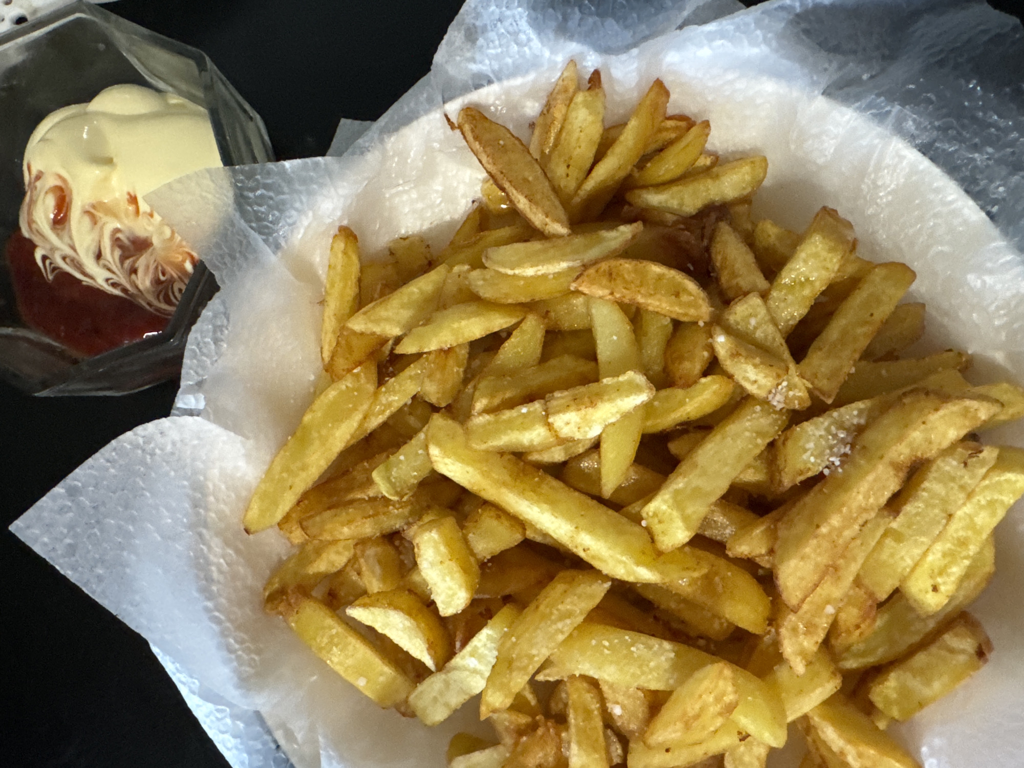 A serving of french fries sits in a white paper-lined container, alongside a clear plastic container holding ketchup and mayonnaise. The fries appear golden-brown and are sprinkled with salt. The ketchup is at the bottom of its container, topped with mayonnaise swirled with more ketchup on top. The location appears to be indoors, and there is no information to indicate it is specifically in Chaikovsky, Russia.