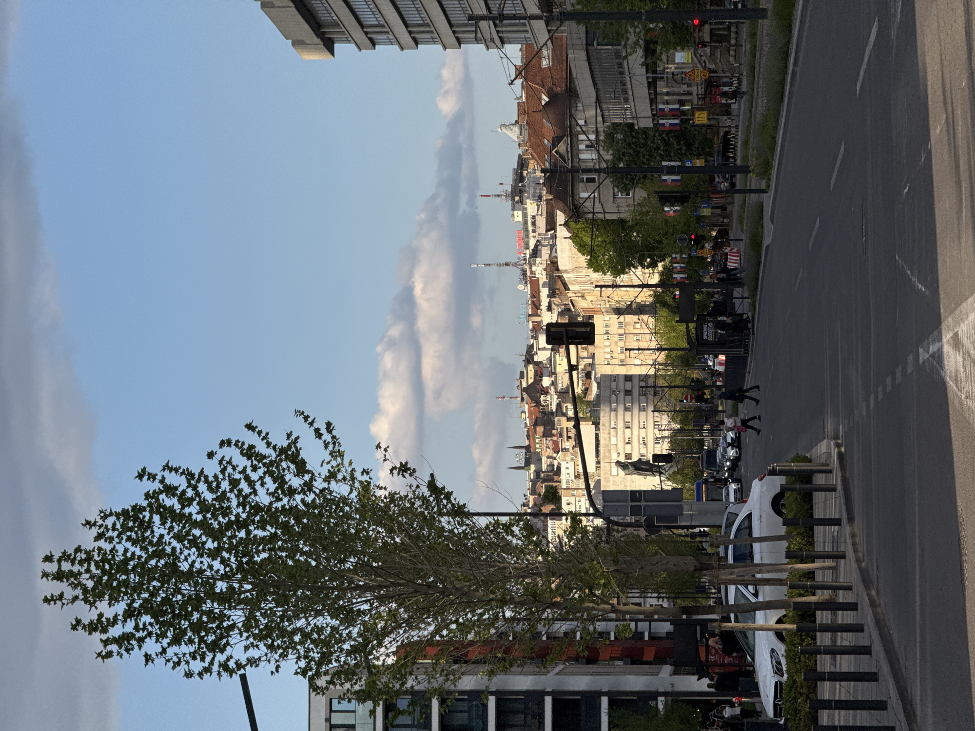 This media file captures an urban street scene in Belgrade, Serbia, depicting a mix of modern and traditional city architecture under a partially cloudy sky. The image appears to be taken in the late afternoon or early evening, indicated by the strong, elongated shadows cast by buildings and objects, and the warm light illuminating some structures.
In the foreground, a leafy tree with bright green foliage stands on the left, partially obscuring a building. To its right, a wide asphalt street with painted lane markings extends into the distance. A sidewalk runs alongside the street, lined with bollards and low shrubbery. Several vehicles, including a prominent white car, are parked along the curb. Pedestrians are visible, with some crossing the street and others walking on the sidewalk.
The mid-ground features a diverse array of buildings, including both contemporary structures and older ones with distinct red-tiled roofs. A statue of a standing figure is visible on the left side of the street. Numerous small Serbian flags, displaying horizontal red, blue, and white stripes, are hung from poles along the right side of the street, confirming the location.
In the background, the cityscape stretches far, showcasing a dense collection of buildings of varying heights, punctuated by several spires and communication towers reaching into the sky. The sky is predominantly blue with scattered white clouds.
Visible text includes a red "GOMEX" sign on a building in the mid-distance and Cyrillic text "СПРЕМАТЕ КУПОВИНУ" (Spremate kupovinu, meaning "Preparing to buy/shop") on a black sign near the street. A yellow diamond-shaped road sign displaying the number "110" is also present.
