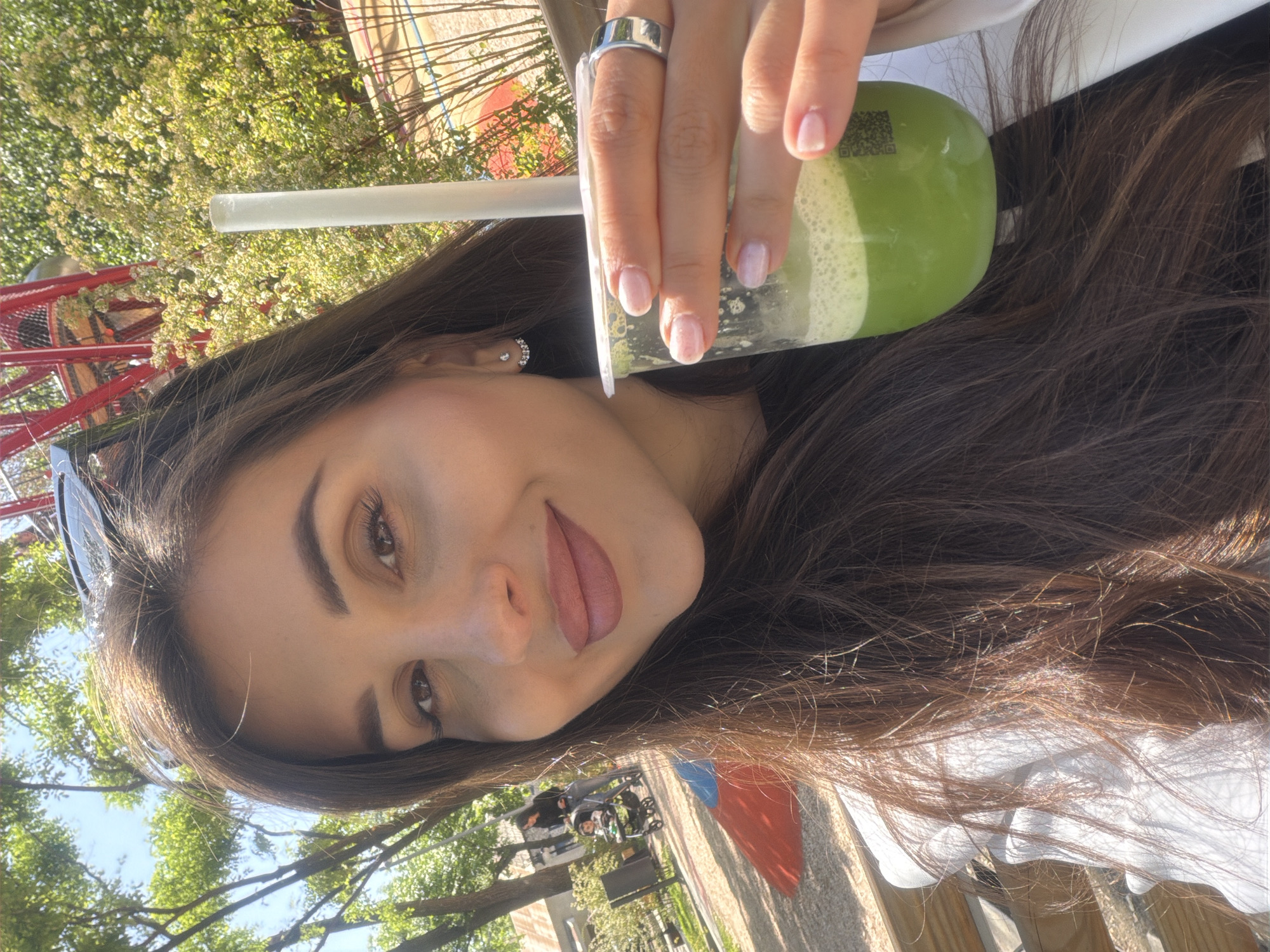 A selfie taken by a woman with long brown hair is shown in a park-like setting. She is holding a cup of a green smoothie, complete with a straw and a white lid. The woman is wearing a silver ring and has light pink nail polish. The background shows trees, a red structure (possibly a carousel), and some people with a stroller, suggesting a public outdoor space. There is no information in the image to identify the exact location as Yerevan, Armenia.