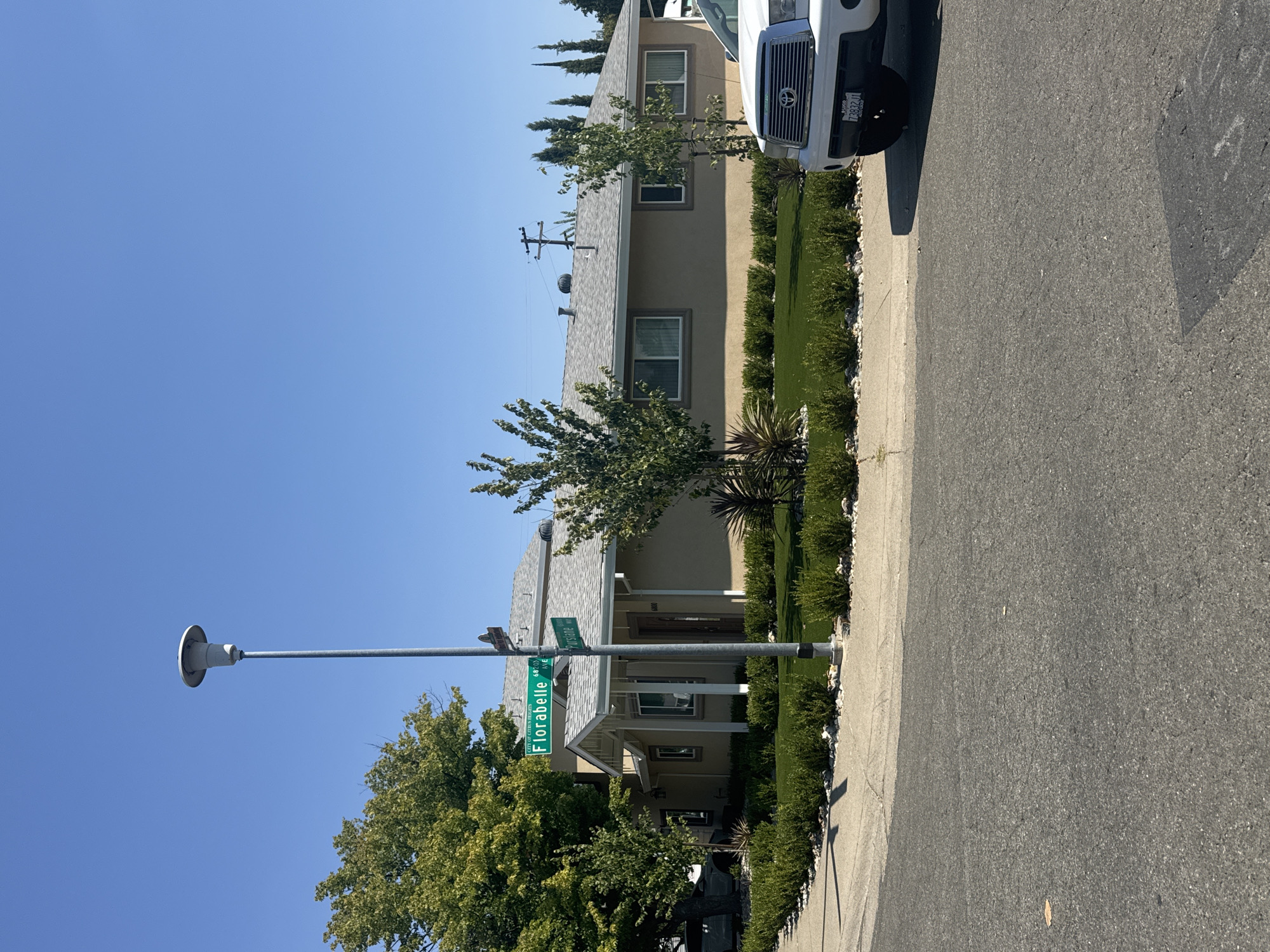 A residential scene is depicted on a clear, sunny day in Citrus Heights, California, USA. The focal point is a light brown, single-story house with a gray shingle roof, located at 6800 Purslane Way. A white pickup truck is parked in front of the house, partially obscuring the driveway. A street sign indicates the intersection of Florabelle Avenue and Purslane Way. The property is landscaped with manicured green bushes and white gravel. A tall street lamp stands near the intersection. The street in the foreground is paved asphalt, with a concrete sidewalk bordering the lawn. There are no people visible in the image.