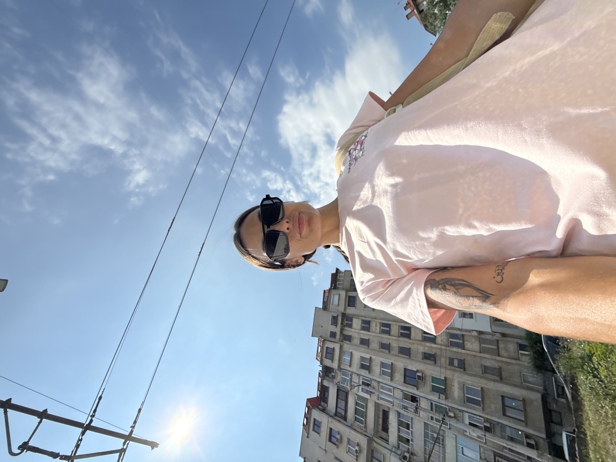 In Pleven, Bulgaria, on a bright, sunny day with a partially cloudy sky, a woman stands outdoors, facing the camera in a low-angle, close-up shot. She is wearing a light pink t-shirt and black sunglasses. Her left arm is visible, displaying a tattoo. Behind her, an old, multi-story apartment building with numerous windows and air conditioning units dominates the background. Overhead, electrical wires crisscross against the sky, suggesting the presence of a tram or trolleybus line. Sunlight is visible on the left side of the image, casting a lens flare. The woman's gaze is directed towards the camera, and her expression is neutral. The overall scene depicts an urban environment on a clear day.