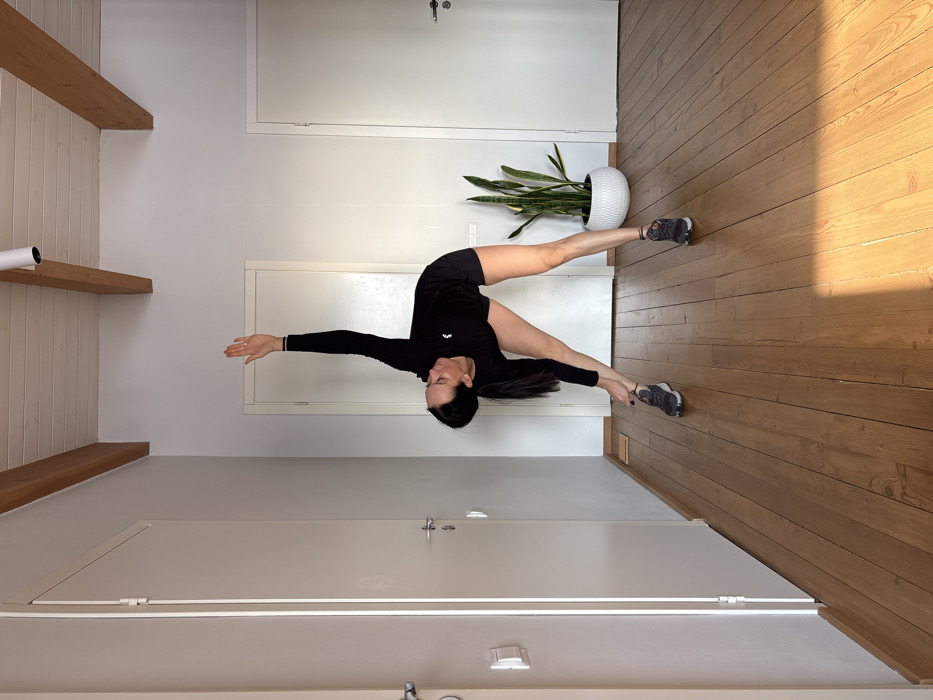 The image depicts an adult female performing a standing side bend or triangle pose variation within an indoor setting in Chaikovsky, Russia. She has dark hair tied back and is dressed in a black long-sleeved athletic top featuring a white logo on the left chest, black athletic shorts, and dark athletic shoes. Her anatomical right hand extends downwards towards her left ankle, while her anatomical left arm is raised vertically, with a ring visible on her left ring finger. The room features light-colored wooden floorboards, partially illuminated by sunlight. White walls line the space, incorporating three white doors with silver-colored handles – one on the left edge and two in the background. A small white electrical outlet is visible on the left wall. The ceiling is white with exposed light-colored wooden beams and a white cylindrical track light. A tall snake plant in a white pot stands on the floor between the two background doors.