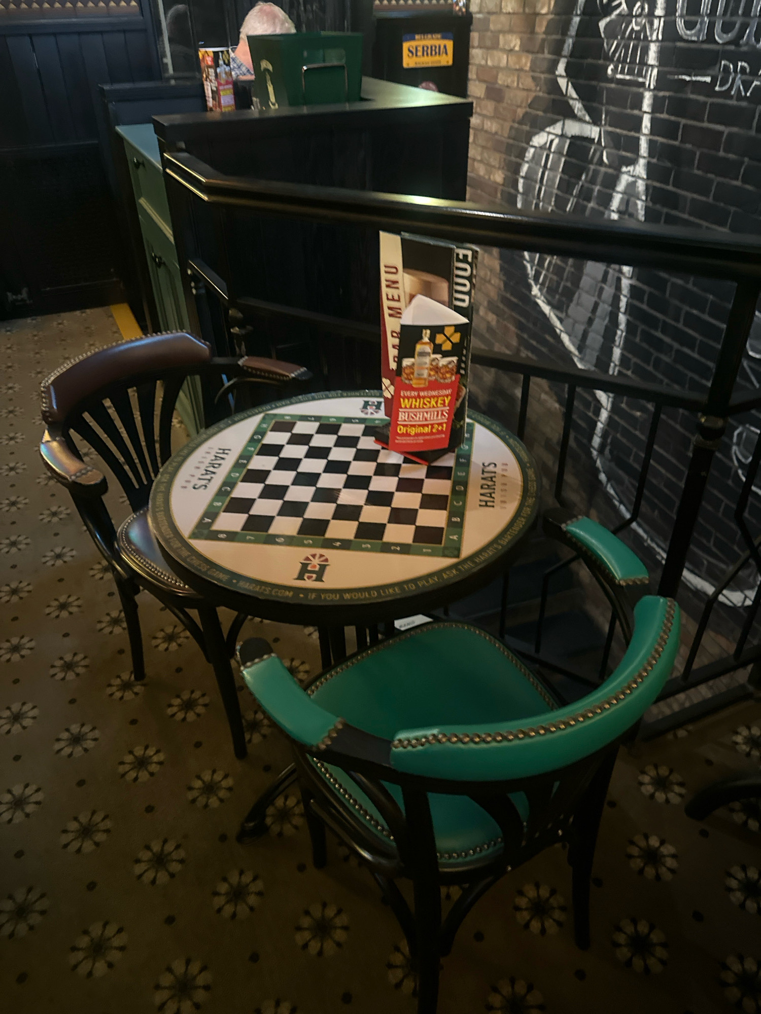 The image shows a small, round table with a chessboard pattern printed on its surface. Two chairs are positioned at the table: one is dark brown with a curved back and wooden slats, and the other is a vibrant green with a curved back and studded trim. A menu stand is placed on the table, displaying a menu that appears to advertise a whiskey promotion. Behind the table, a dark metal railing separates the area from a brick wall with white chalk drawings. A small yellow sign with the word "SERBIA" is visible on a dark structure to the left of the scene, suggesting the location is in Belgrade, Serbia. The floor is covered in a patterned carpet with floral designs and small dark dots. The overall lighting is dim, creating a moody atmosphere. No people are clearly visible in the frame, but a partial view of a person with gray hair can be seen in the background, behind the counter area.