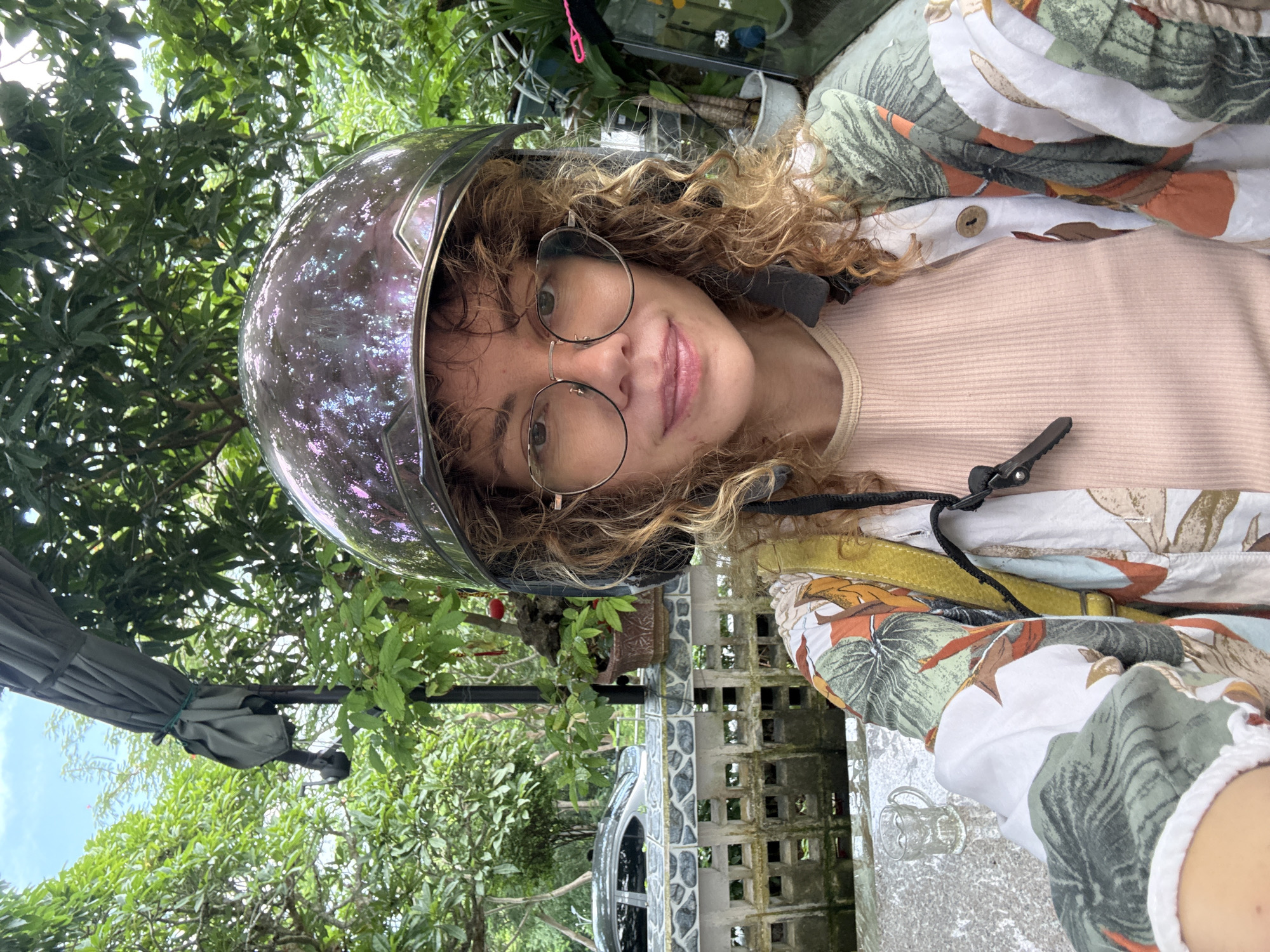A selfie shows a woman in a motorcycle helmet and glasses, wearing a light pink top and a brightly patterned shirt. She is outdoors, with lush green foliage and trees in the background, suggesting a tropical or garden setting. A partially visible grey car is parked behind a decorative fence. The scene appears to be in Ho Chi Minh City, Vietnam, a vibrant metropolis known for its bustling streets and abundant greenery.