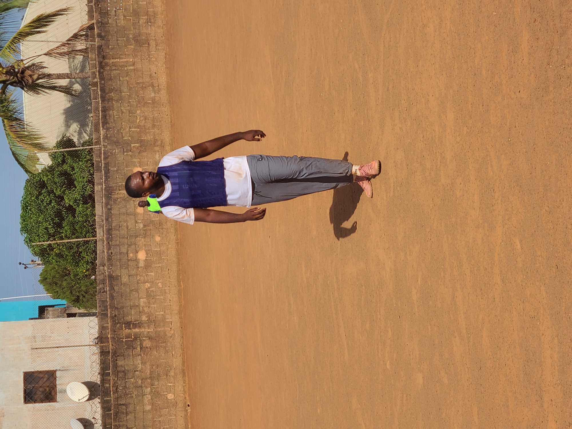 A man lies on his back on a dirt surface, possibly a sports field. He wears a white shirt, blue vest, gray pants, and pink shoes. His right arm is extended above his head, and his left arm is extended downwards. A neon green object is near his neck. The background shows a brick wall, trees, and a building in the distance, partially obscured by a chain-link fence. A palm tree is visible in the upper left. The location is suggested to be Barakin Lerawi, Nigeria.
