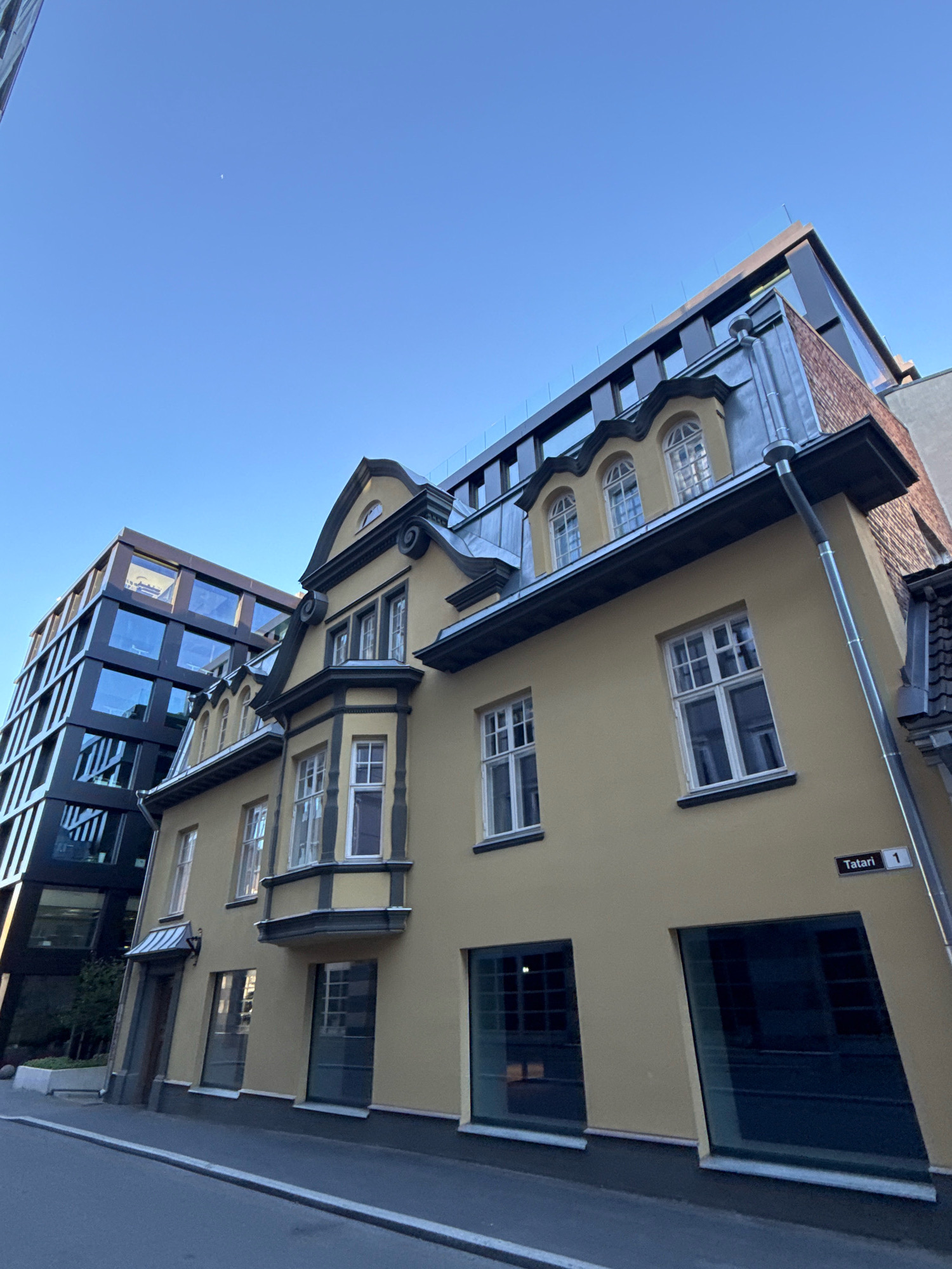 The image depicts a street scene in Tallinn, Estonia, featuring two distinct buildings. The foreground is dominated by a yellow building with traditional architectural elements, including arched dormer windows with decorative scrollwork, and a bay window. This building has a gray roof and a visible gutter system. To the left of the yellow building is a modern, dark glass building with a geometric design and multiple stories. The sky above is clear and blue, with a small, faint crescent moon visible. A street sign reading "Tatari 1" is affixed to the wall of the yellow building. The street in front of the buildings appears to be paved with asphalt and has a white line marking. The scene is captured from a low angle, looking upwards. No people or animals are visible in the image.