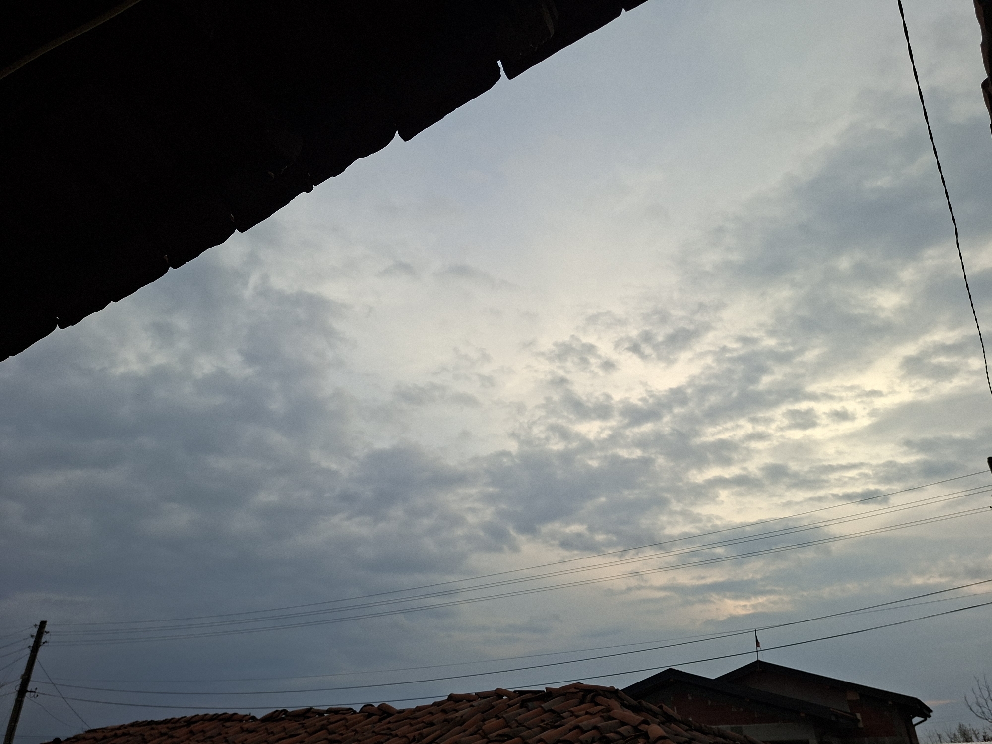 The image displays an upward view of an outdoor scene within a residential area of Zhitnitsa, Bulgaria, under an overcast sky with diffused light. A dark, silhouetted architectural element, likely a roof eave, partially obscures the upper left portion of the frame, with a thin, dark vertical wire extending downward from the upper right edge. The sky is dominated by a formation of grey and light-toned altocumulus clouds. Multiple parallel utility wires traverse horizontally across the mid-ground. In the lower left quadrant, a wooden utility pole equipped with insulators is visible. The lower section of the image features the upper parts of several structures, including a prominent roof composed of reddish-brown terracotta tiles. Adjacent to this, a building with a darker-toned roof and upper wall is visible, distinguished by a small, dark flag positioned at its apex. No individuals or specific actions are present in the depicted scene.