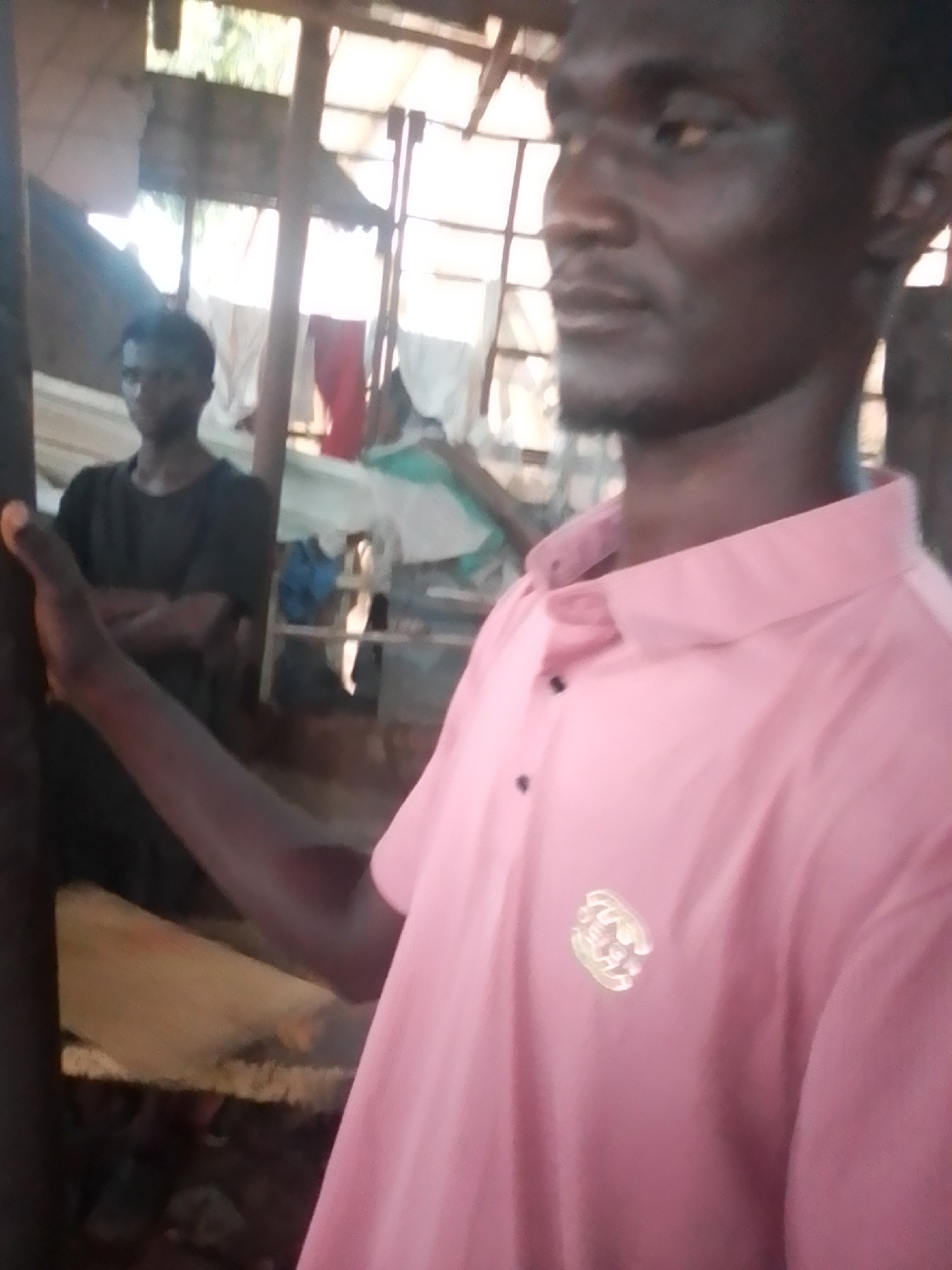 In Pantame, Nigeria, a man in a pink collared shirt with a circular emblem on the left chest is captured in a medium close-up shot, facing slightly to his left. His dark skin, short dark hair, and a thin beard are visible. In the blurred background, another man wearing a dark t-shirt stands with his arms crossed. The environment appears to be an outdoor or semi-outdoor setting, possibly a market or workshop, with a wooden structure overhead and various items suggesting storage or goods for sale. The lighting is natural and somewhat diffuse, creating a slightly grainy image quality.
