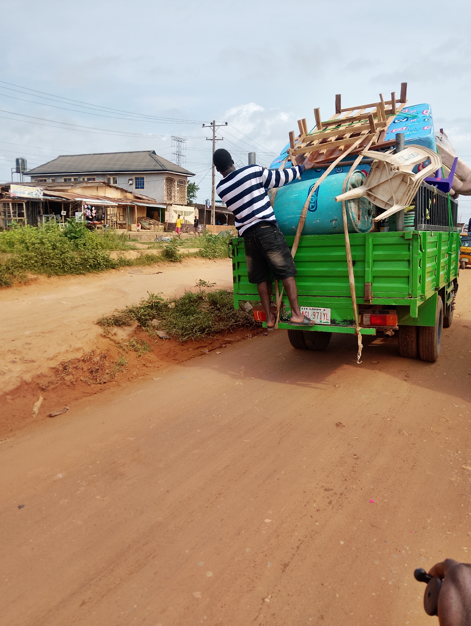 A man in a black and white striped shirt and black shorts is standing on the back of a green truck laden with various items. He is holding onto a large blue object and appears to be adjusting it. The truck is parked on a dirt road in a city. In the background, there are buildings, some of which appear to be shops. There are also power lines and a transmission tower visible. The sky is overcast. The license plate on the back of the truck reads "LAGOS AGL 971YL". This scene is located in Agege, Nigeria.